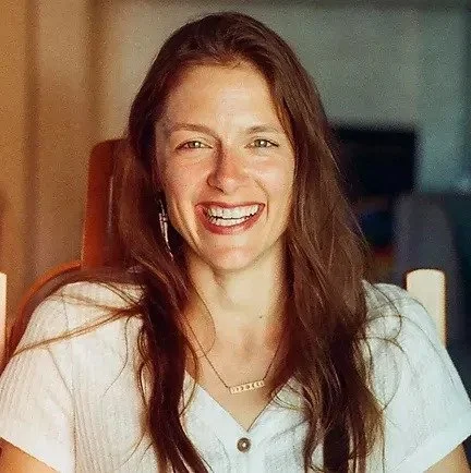 A smiling woman with long brown hair, wearing a light-colored top and jewelry, in a warmly lit indoor setting.