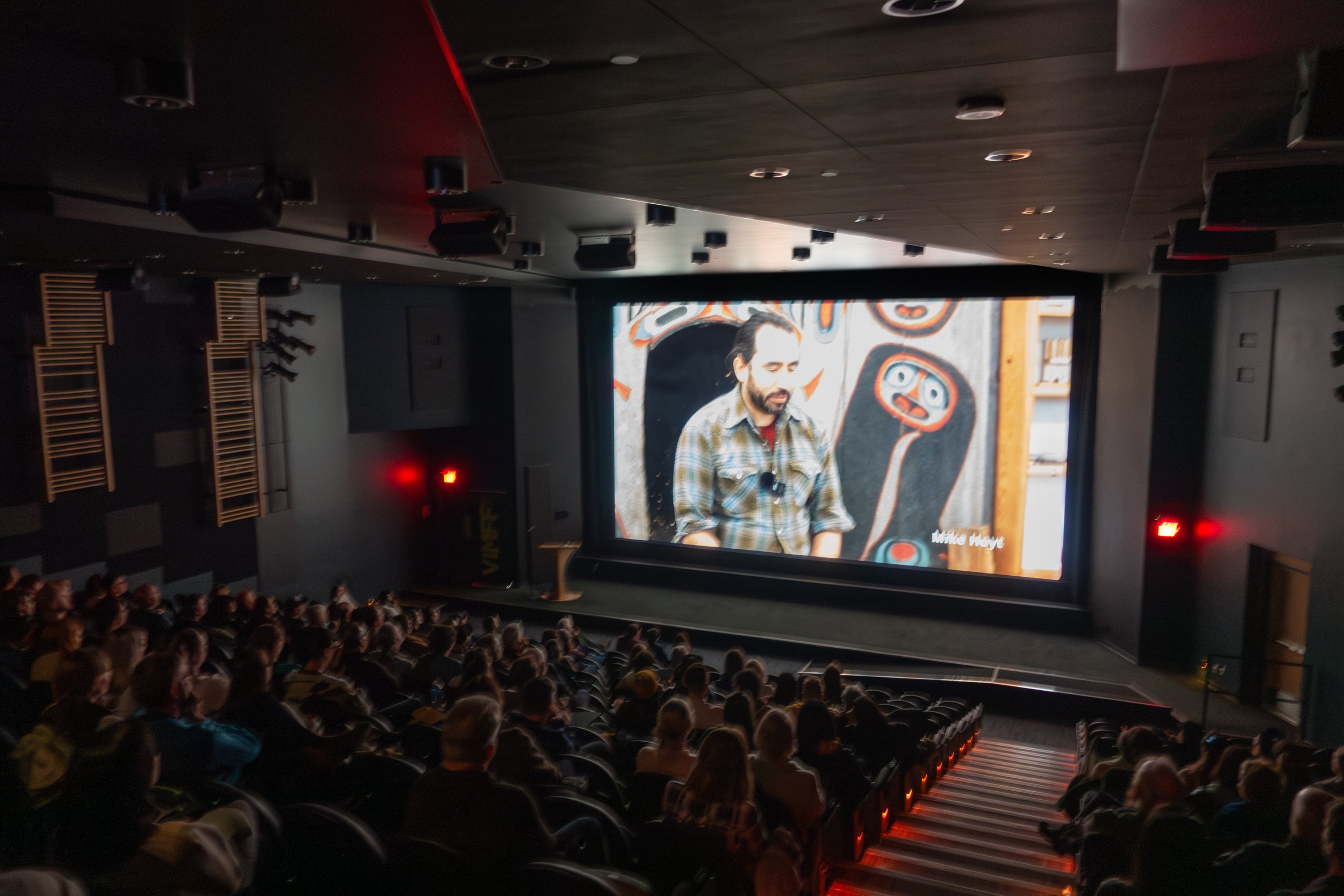 Audience watching a presentation or film in a dark theater with a large screen showing a man with a beard and plaid shirt speaking, and background artwork.