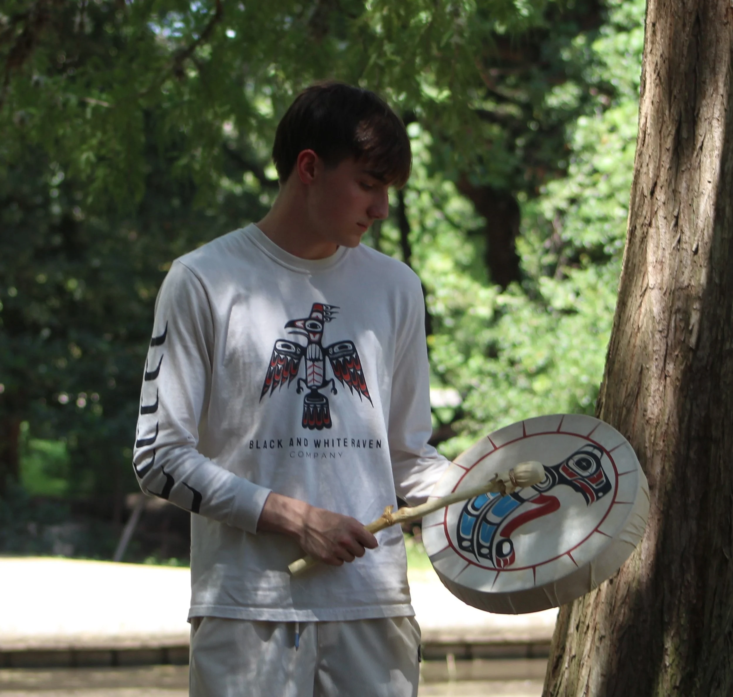 A young man holding a traditional Native American hand drum, standing outdoors next to a tree, with lush green foliage in the background.