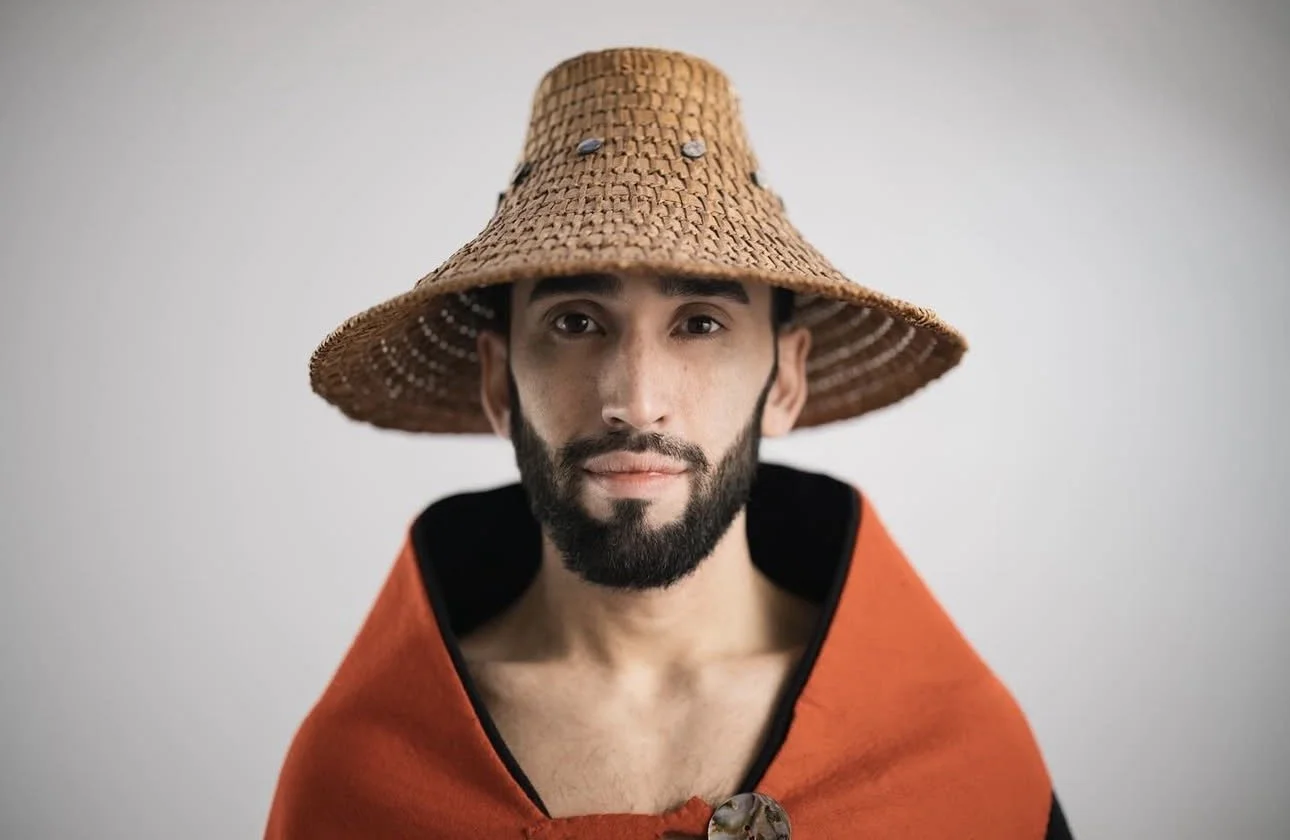 A man with a beard wearing a large woven straw hat and an orange cape with black trim, standing against a plain light backdrop.