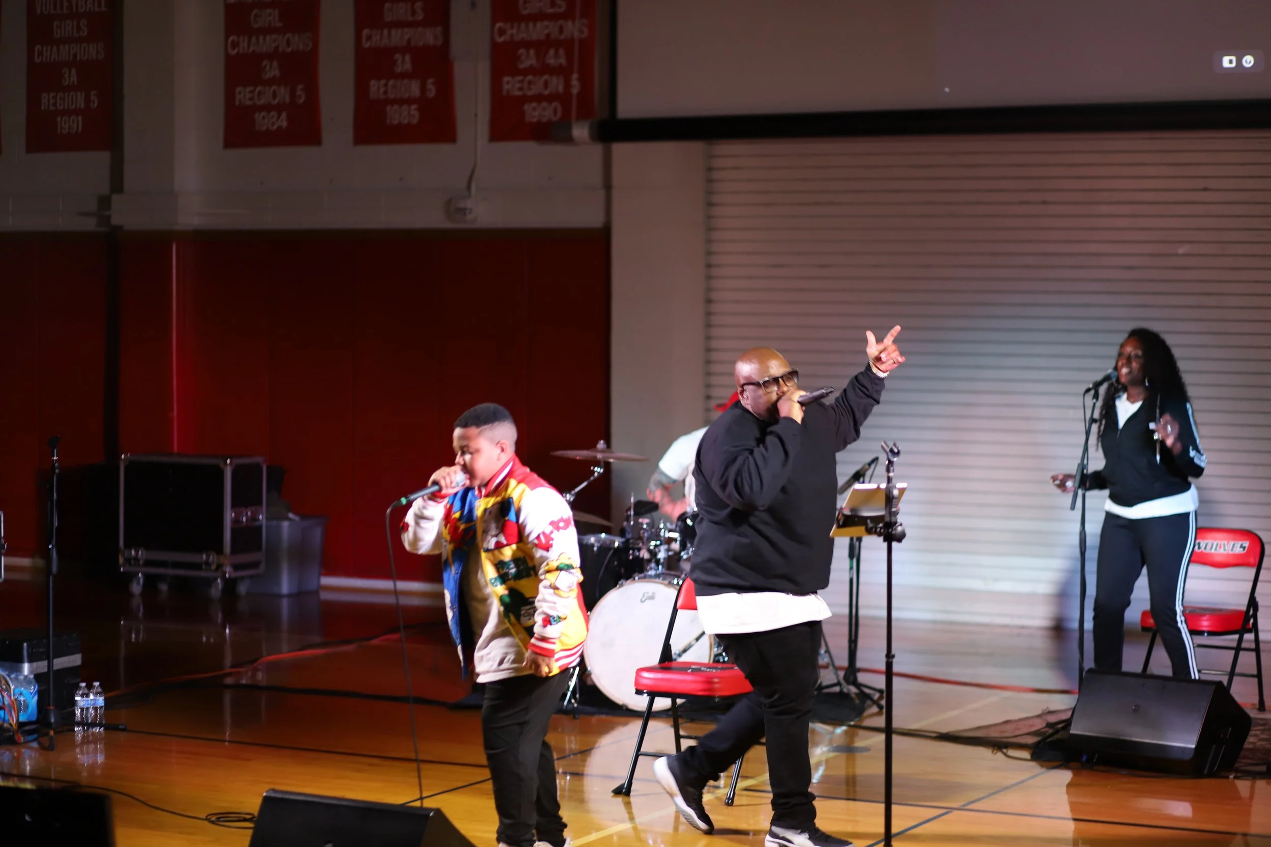 Performance scene on stage with two singers, a man and a boy, holding microphones, a woman singing backup, and a drummer in the background, with banners hanging on the wall and musical equipment on stage.