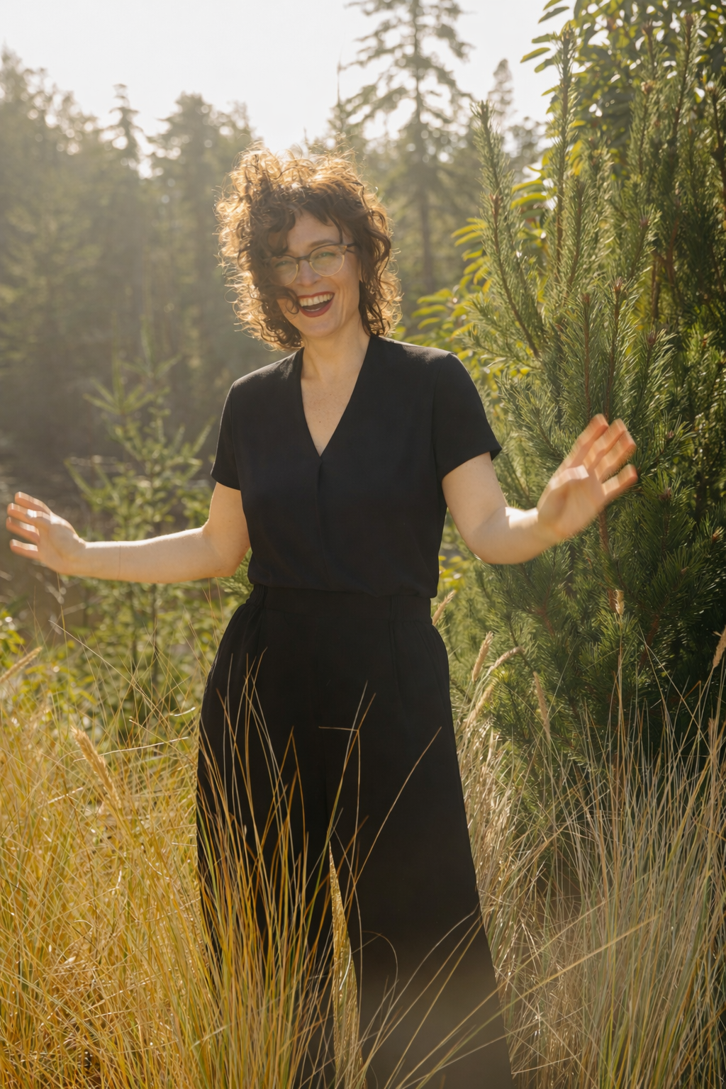 A woman with curly hair and glasses, smiling and wearing a black outfit, standing outdoors in a field of tall grass with green trees in the background.