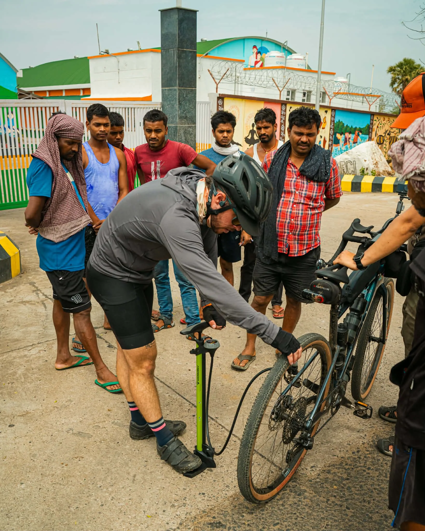 Cyclist adjusting a bike wheel for a group of onlookers outside near a colorful building and mural.