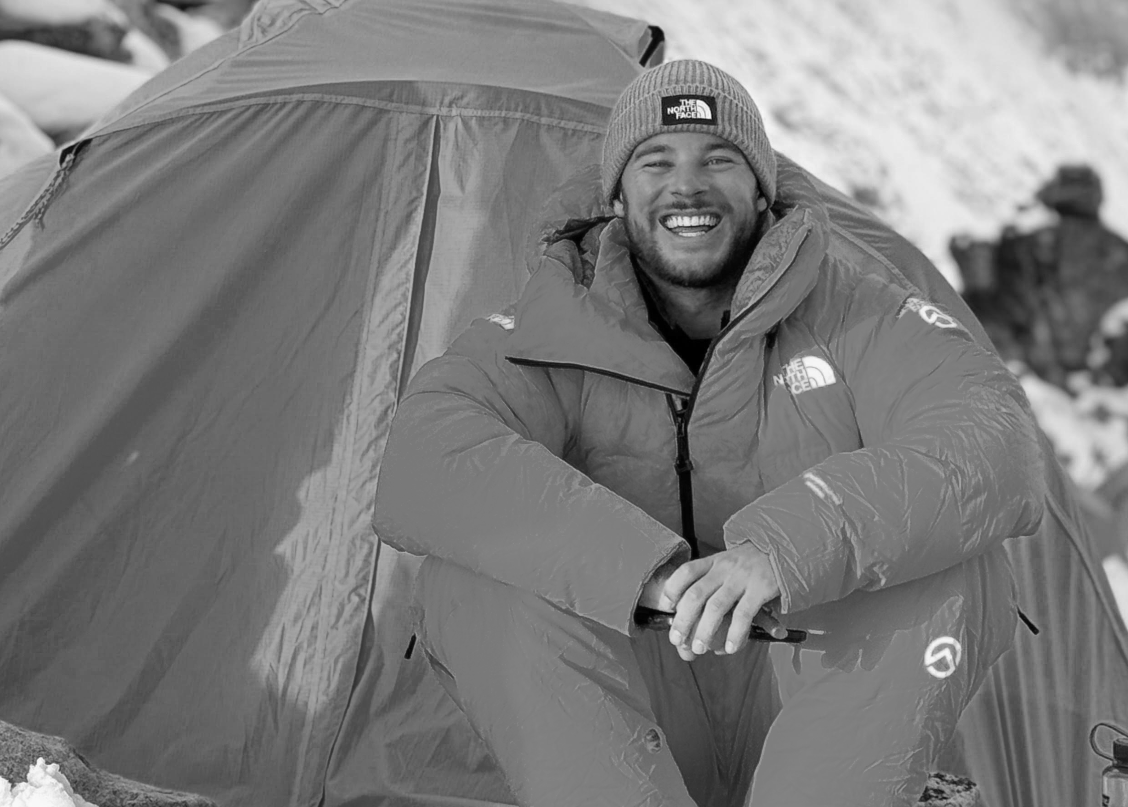 Smiling man in outdoor winter gear sitting near a tent in a snowy landscape, wearing a beanie and a North Face jacket.