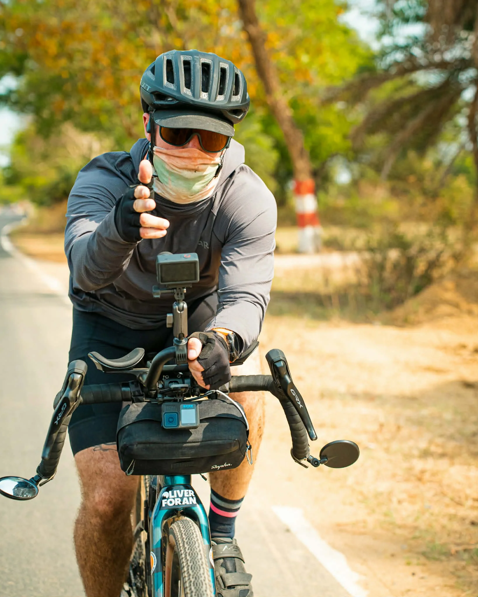 A man biking outdoors wearing a helmet, sunglasses, a face mask, gloves, and a long-sleeve shirt, pointing toward the camera.