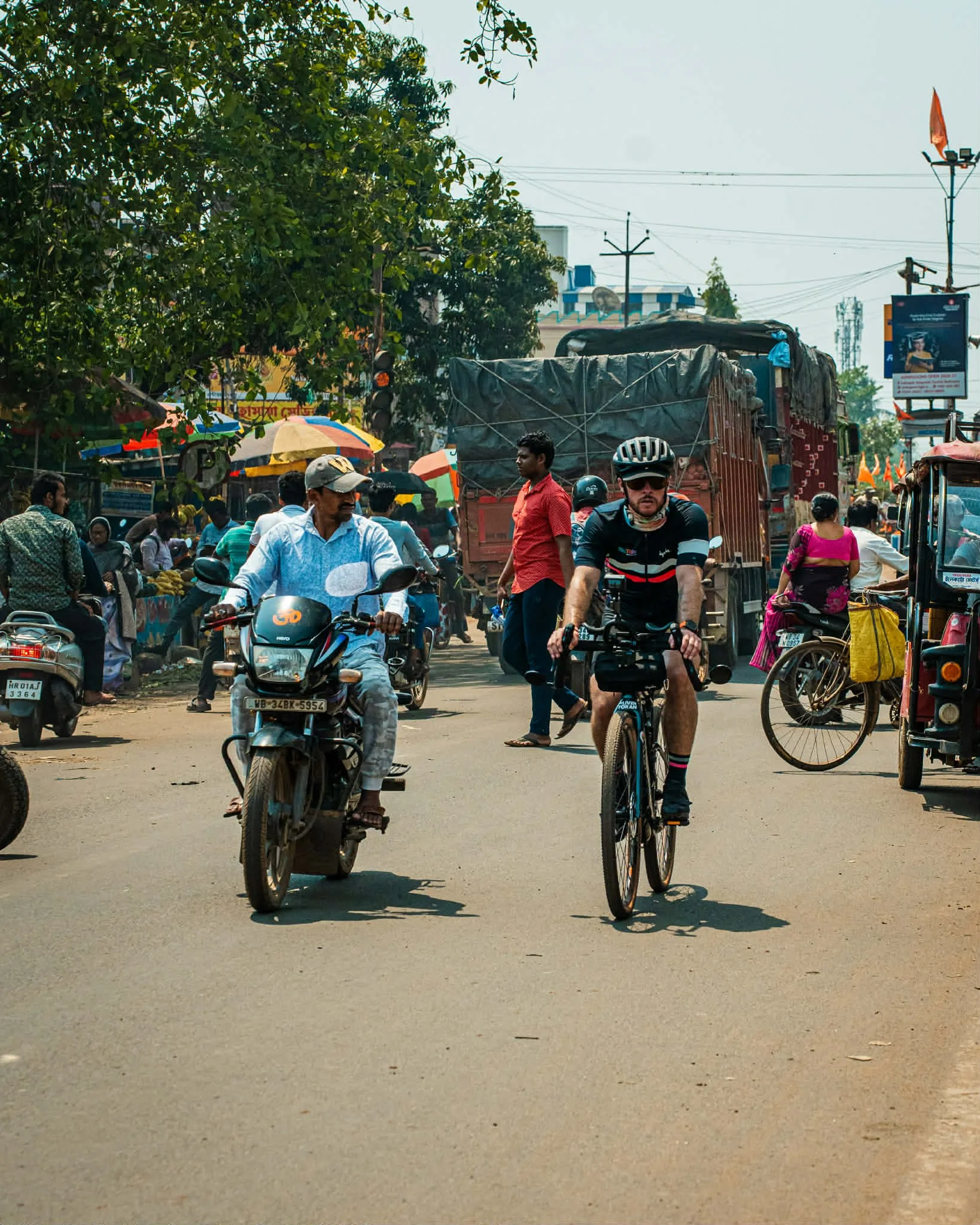 A busy street scene with people riding bikes and motorcycles, and various other vehicles and pedestrians in the background, under a clear sky.