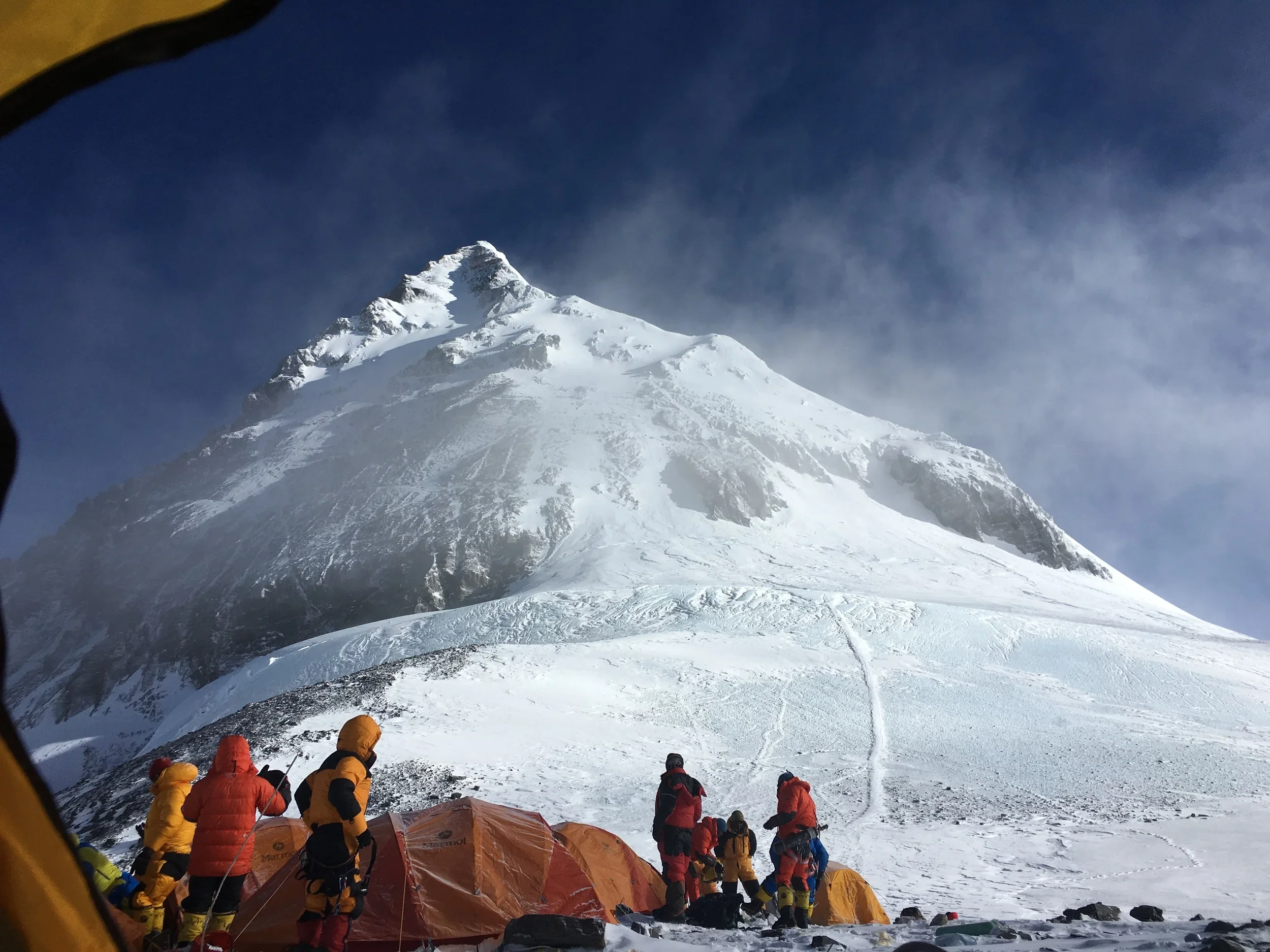 Climbers in colorful gear camping near a snow-covered mountain with a steep peak, under a blue sky with some clouds.