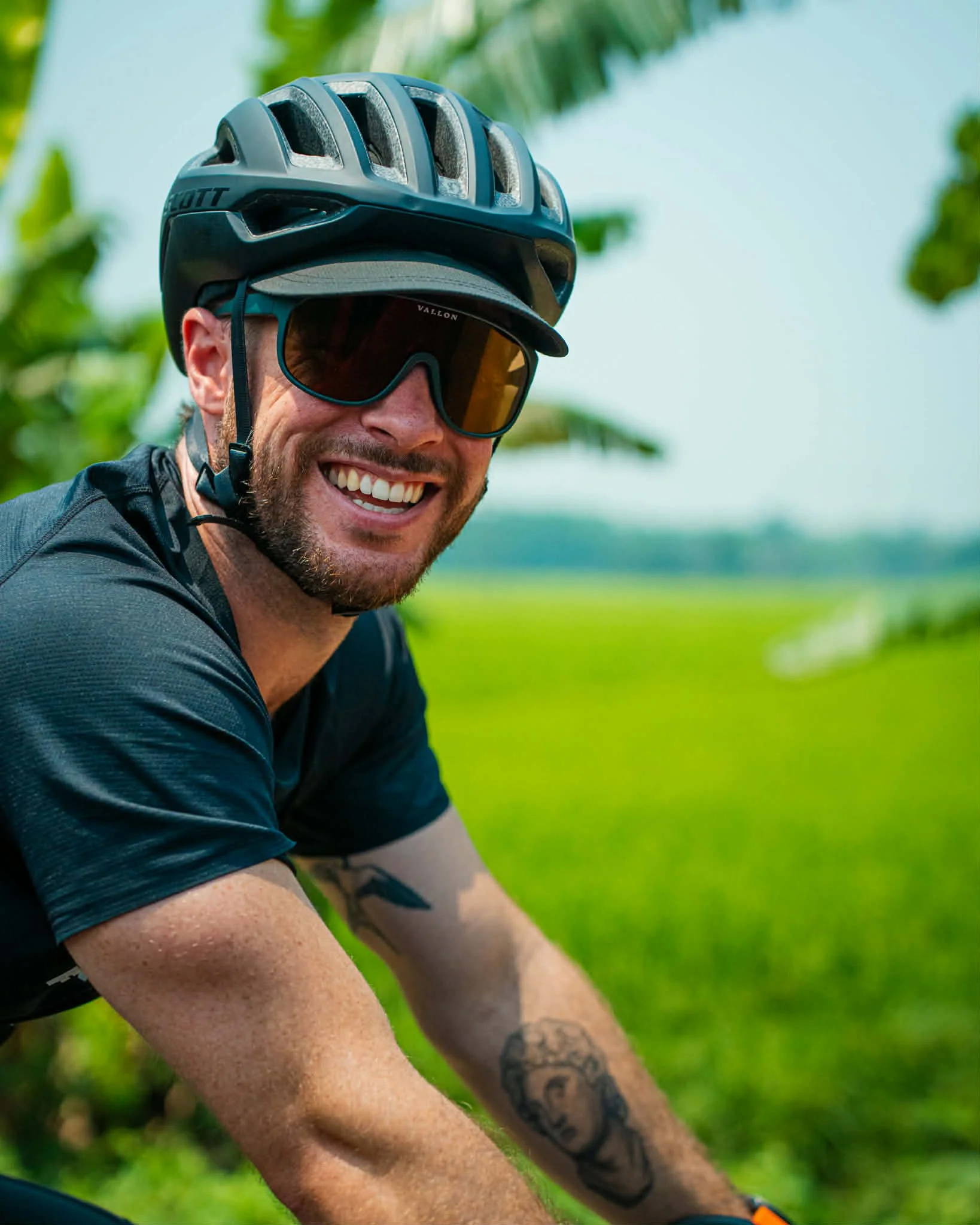 A smiling man wearing a bike helmet, sunglasses, a black shirt, and tattoos on his arms, outdoors in a lush green environment.