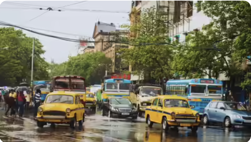 Street scene with yellow taxis, modern cars, buses, pedestrians, wet pavement, trees, and buildings in the background.