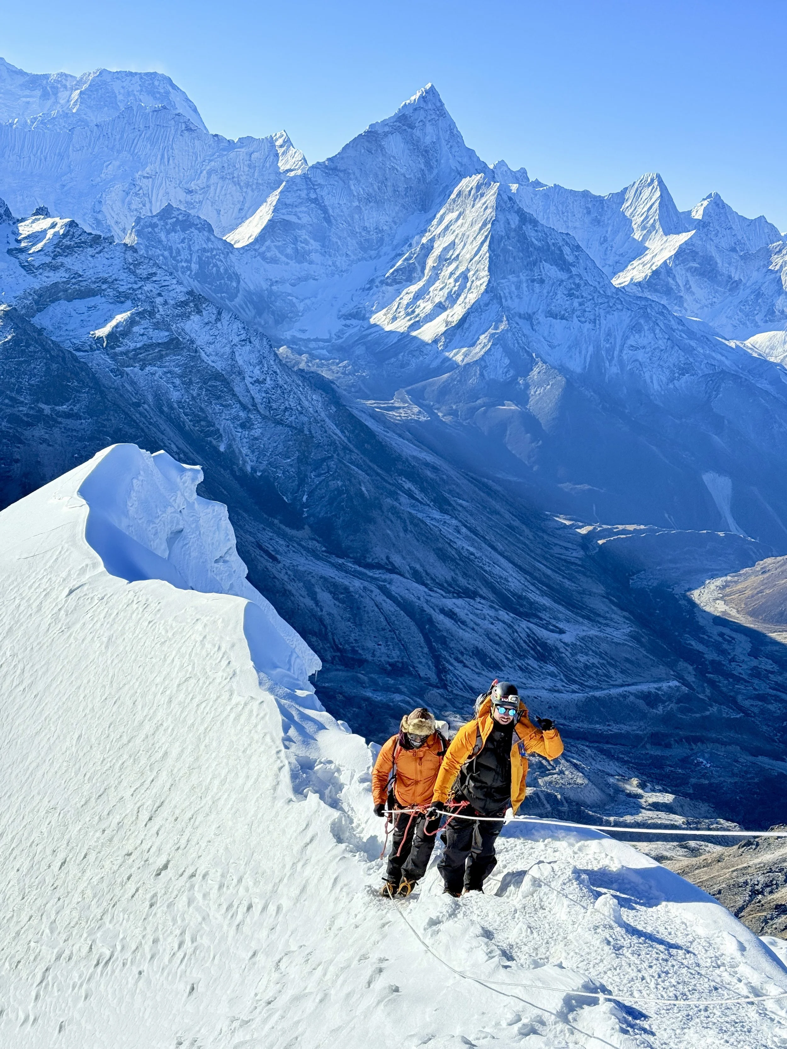 Two climbers in orange and black gear ascending a snowy mountain ridge with tall, snowy peaks in the background.