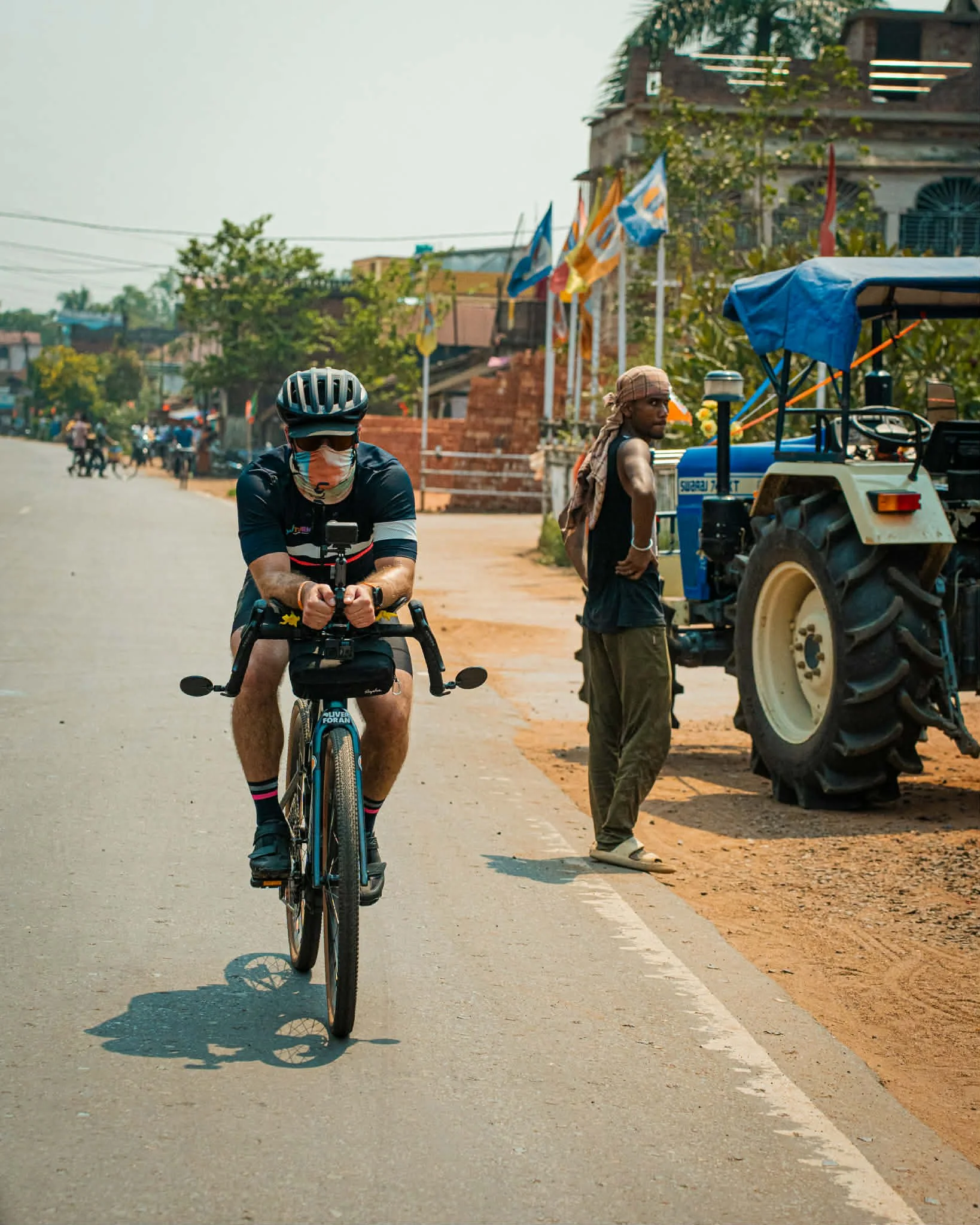 A man riding a bicycle on a dusty street in a rural or semi-urban area, wearing a helmet and face mask, with a person standing near a tractor on the side of the road, and flags flying in the background.