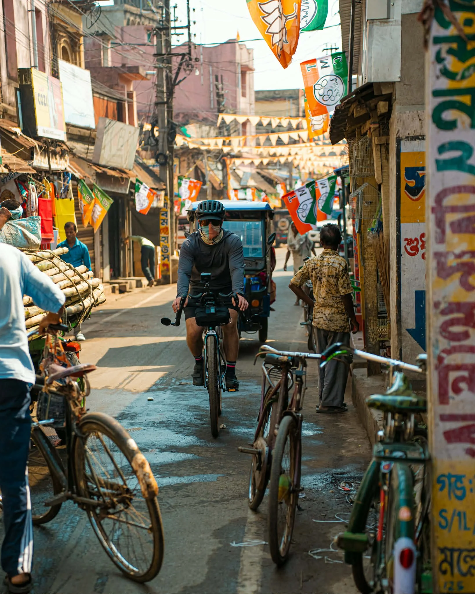 A busy street scene in an urban area with multiple people, bicycles, and a tuk-tuk. Flags and banners hang above the street, and buildings with small shops line the sides. A man wearing a helmet and sunglasses is riding a bicycle, while other pedestr