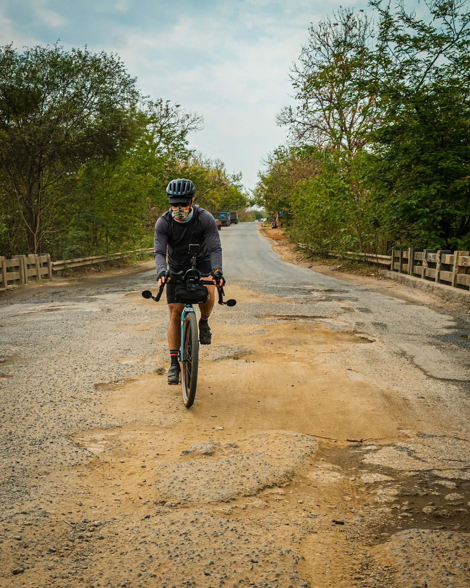 A cyclist rides on a rough, pothole-filled road with green trees on both sides.