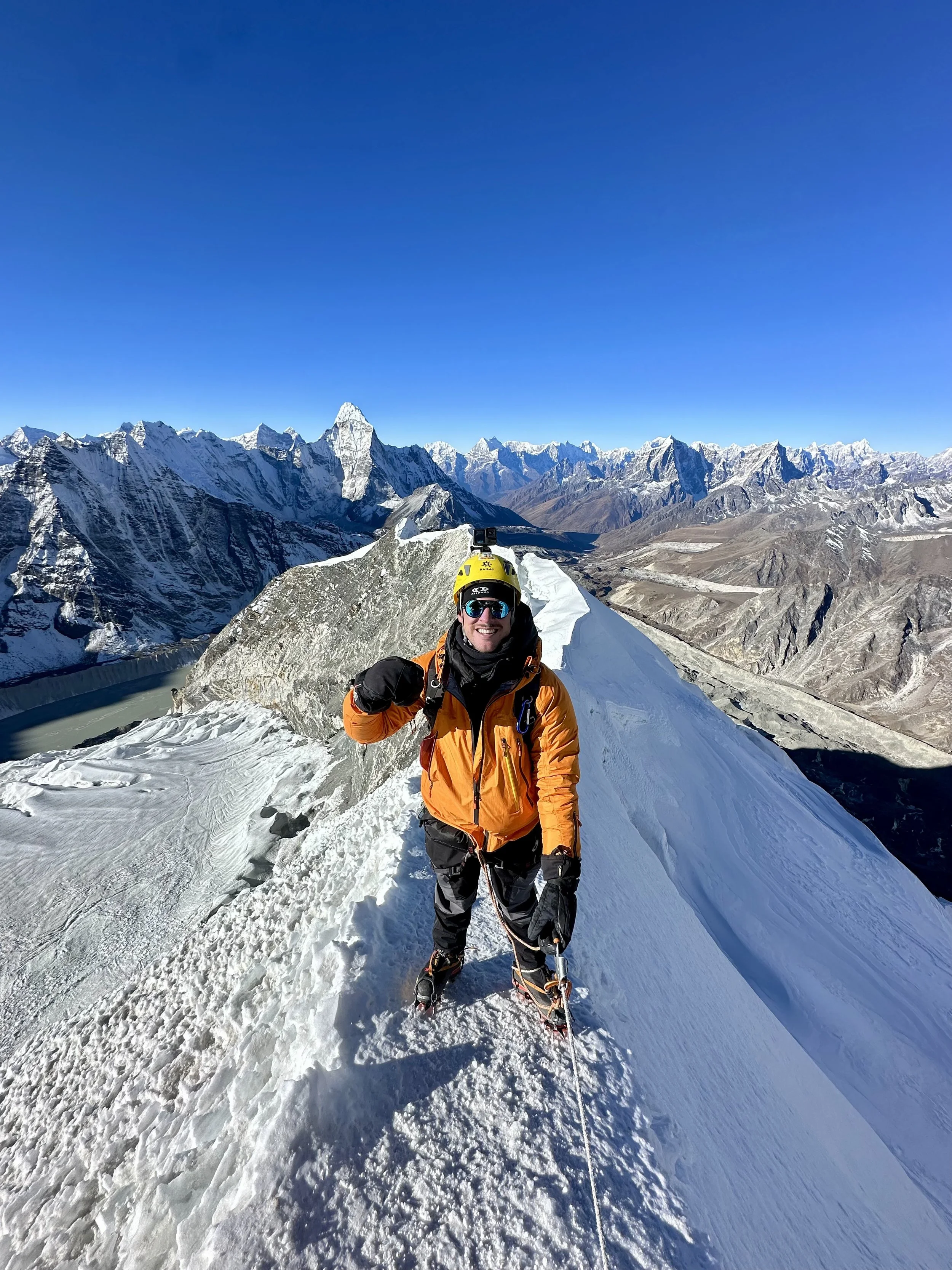 Climber standing on a snowy mountain ridge, smiling with thumbs up, wearing a yellow helmet and orange jacket, surrounded by snow-covered peaks and blue sky.