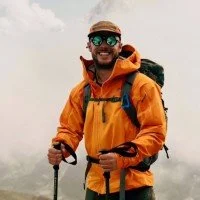 Smiling man in orange outdoor jacket with sunglasses and hat, holding trekking poles, against a foggy background.
