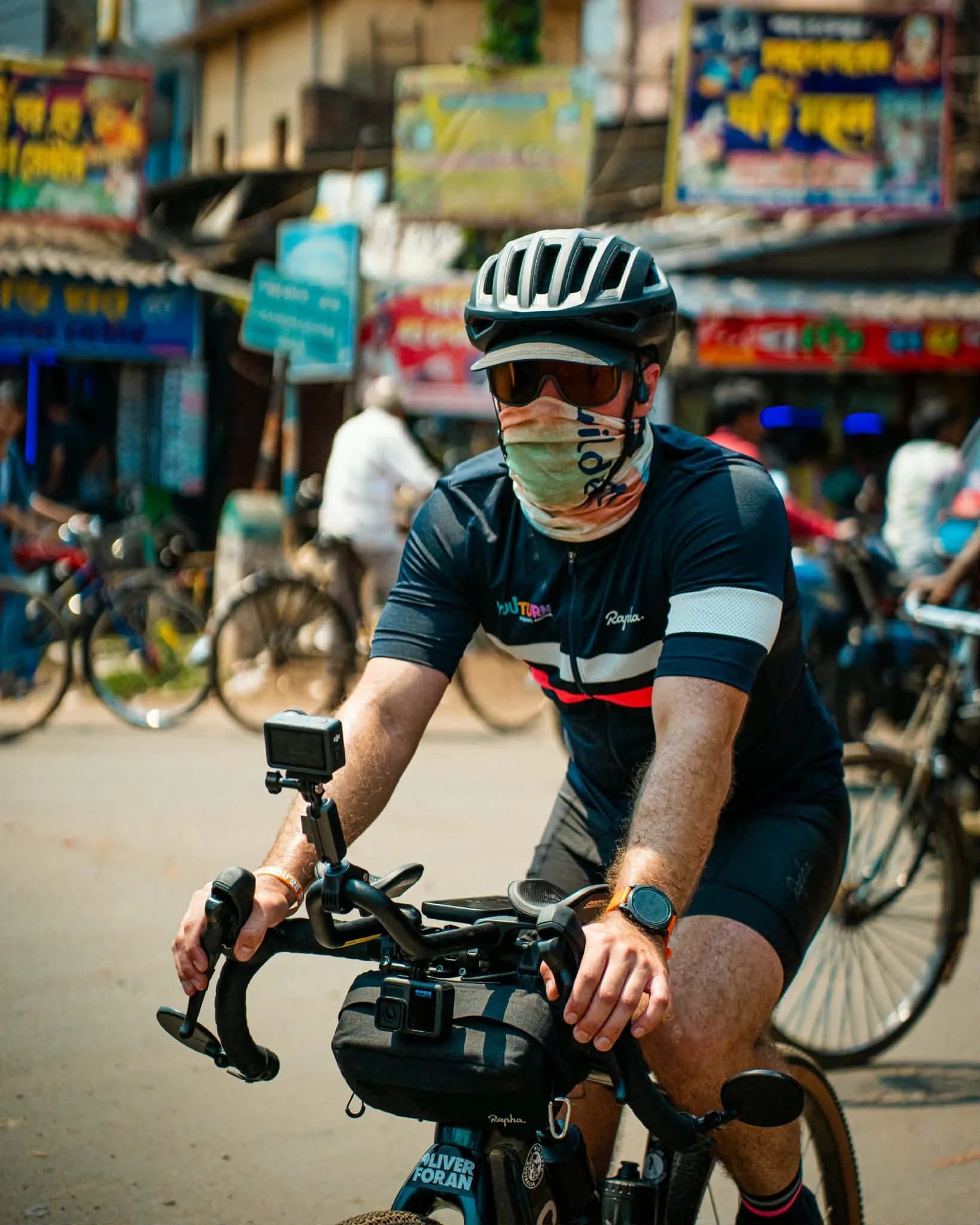 Man riding a bicycle on a busy street, wearing a helmet, sunglasses, a face covering, and a sports watch, with shops and other cyclists in the background.
