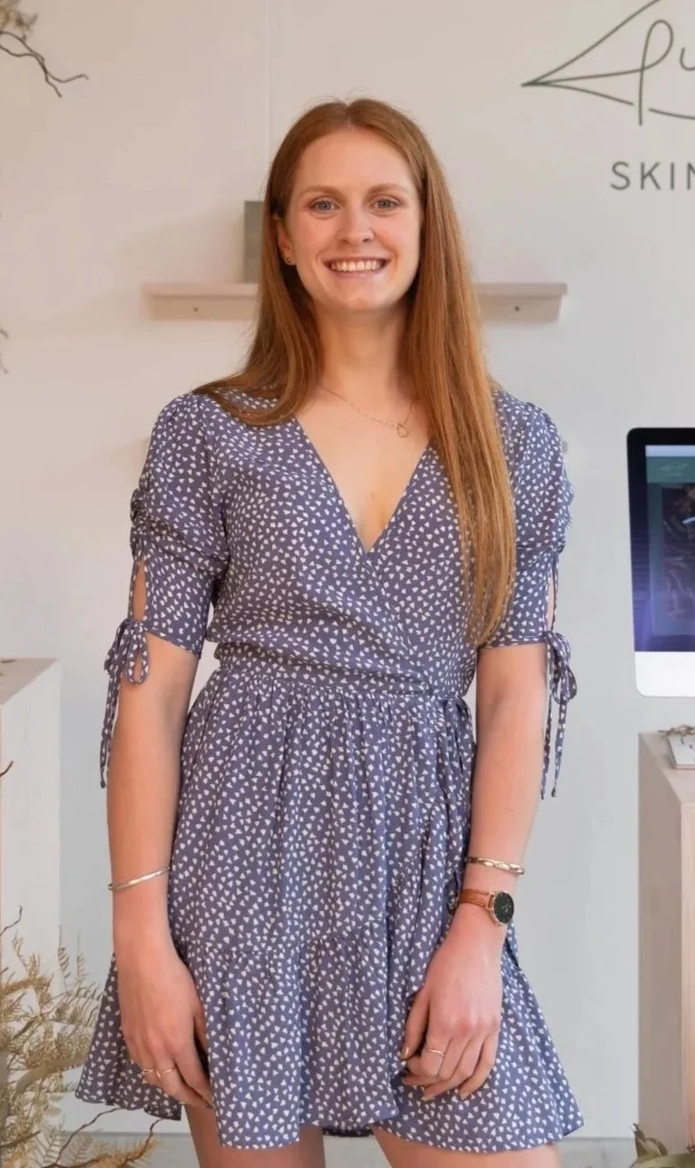A woman with long red hair wearing a blue dress with white small triangle patterns, standing inside a room with a white wall, a computer monitor, and some decorative plants.