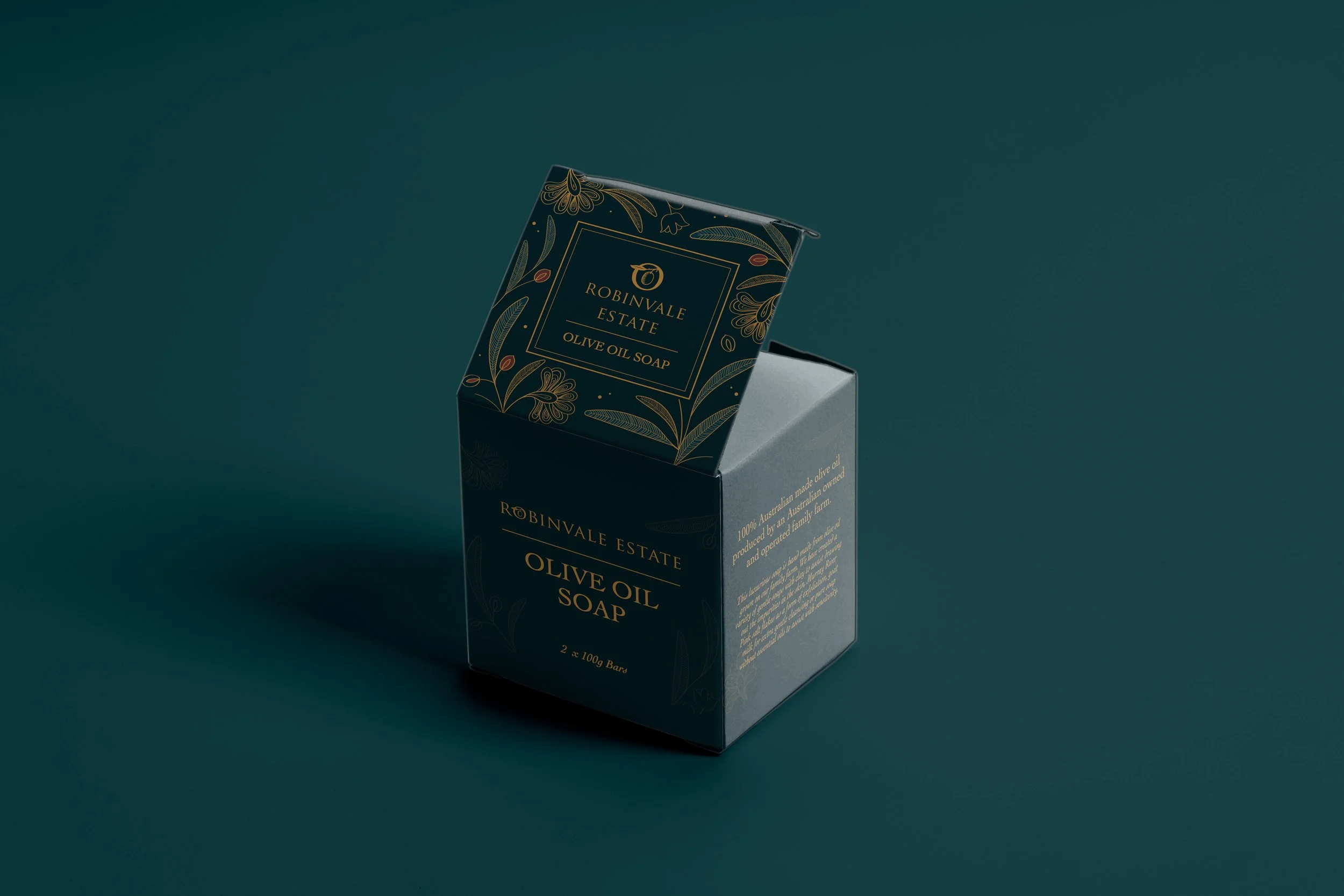 A dark blue box of Robinvale Estate olive oil soap with gold floral accents, open to reveal two soap bars inside, placed on a matching dark blue surface.