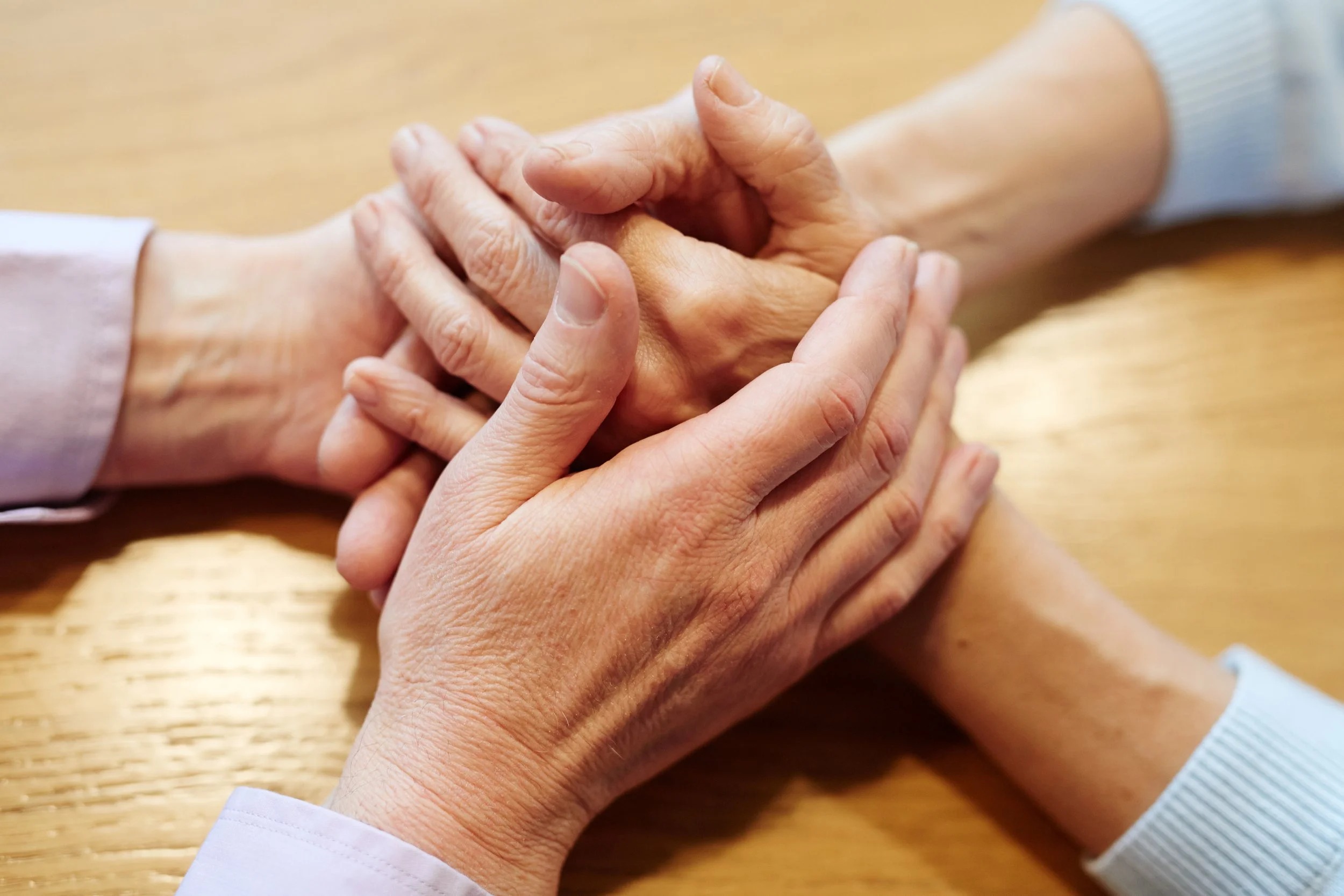 Two people are holding hands, one appears to be an elderly person and the other an adult, on a wooden surface.