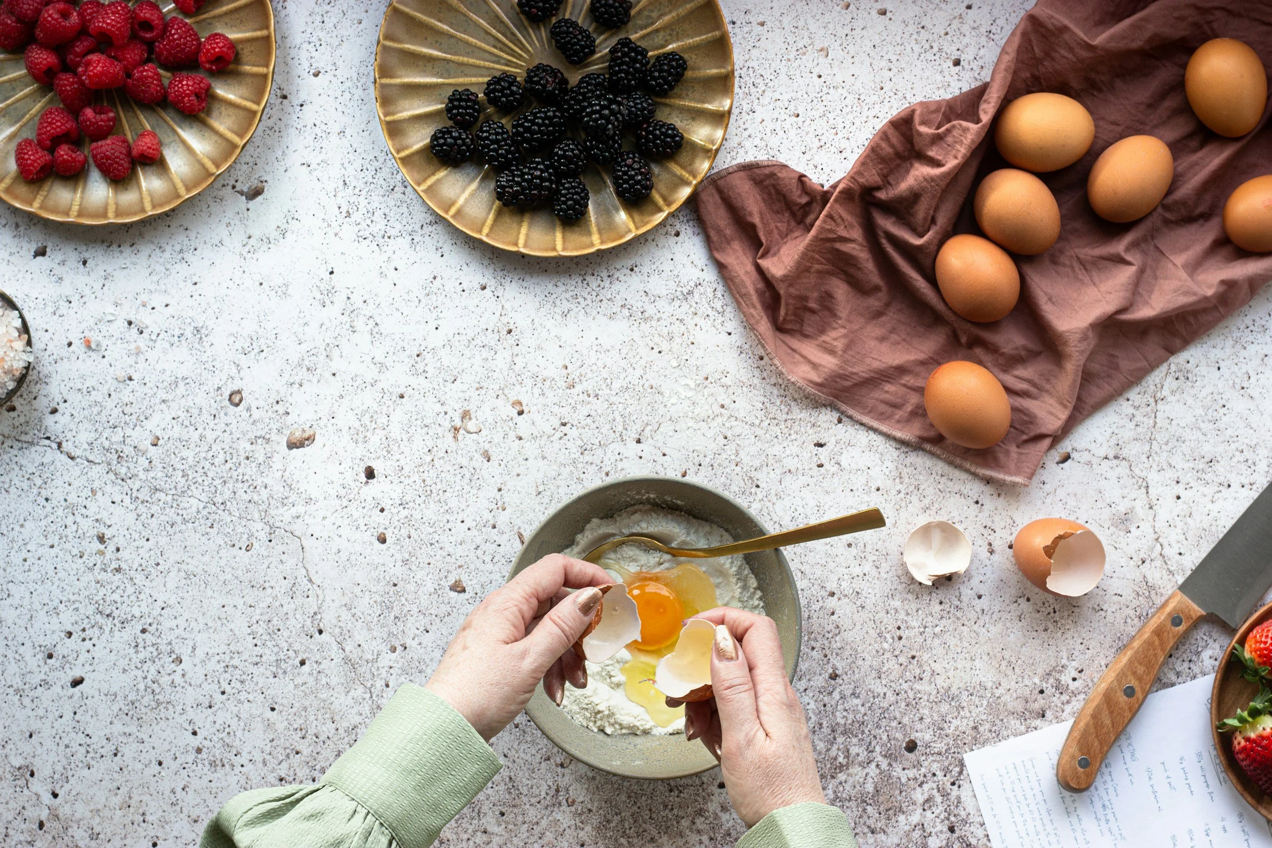 A person cracking eggs into a bowl of flour with berries and eggs on the counter.