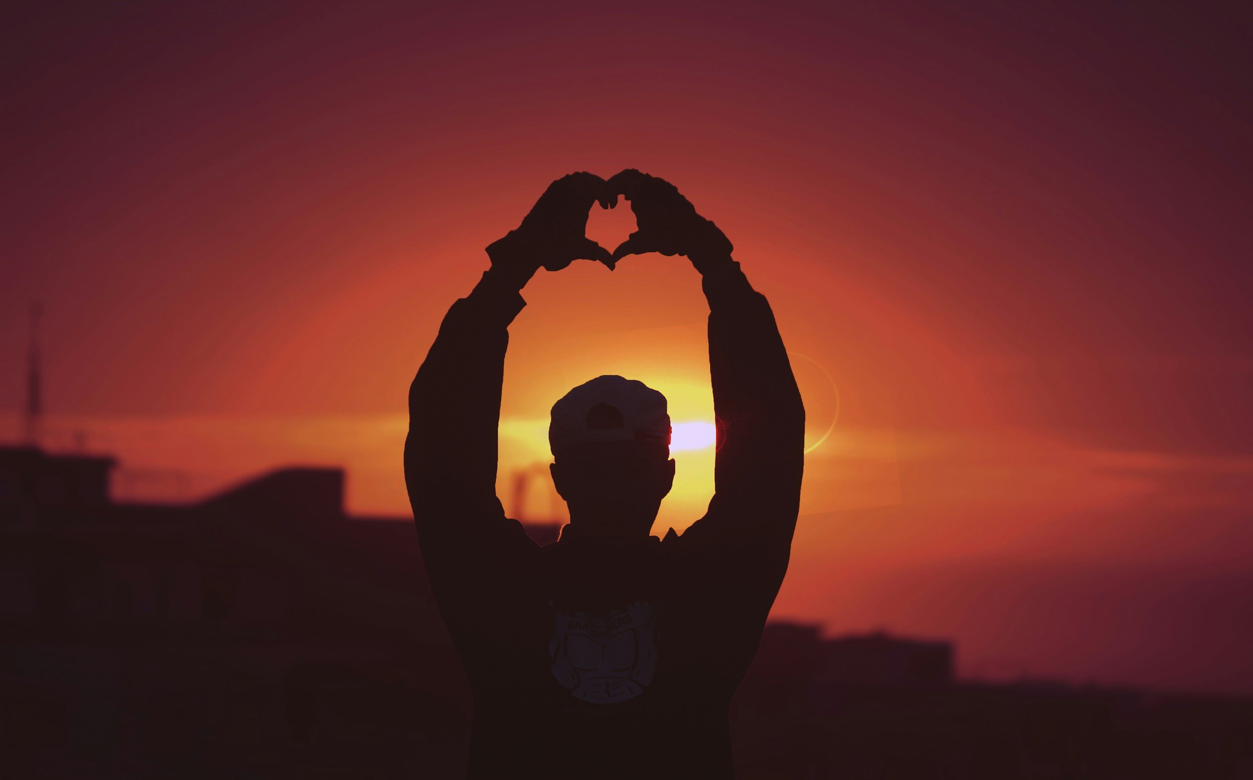 Person silhouetted against a sunset with hands forming a heart shape.