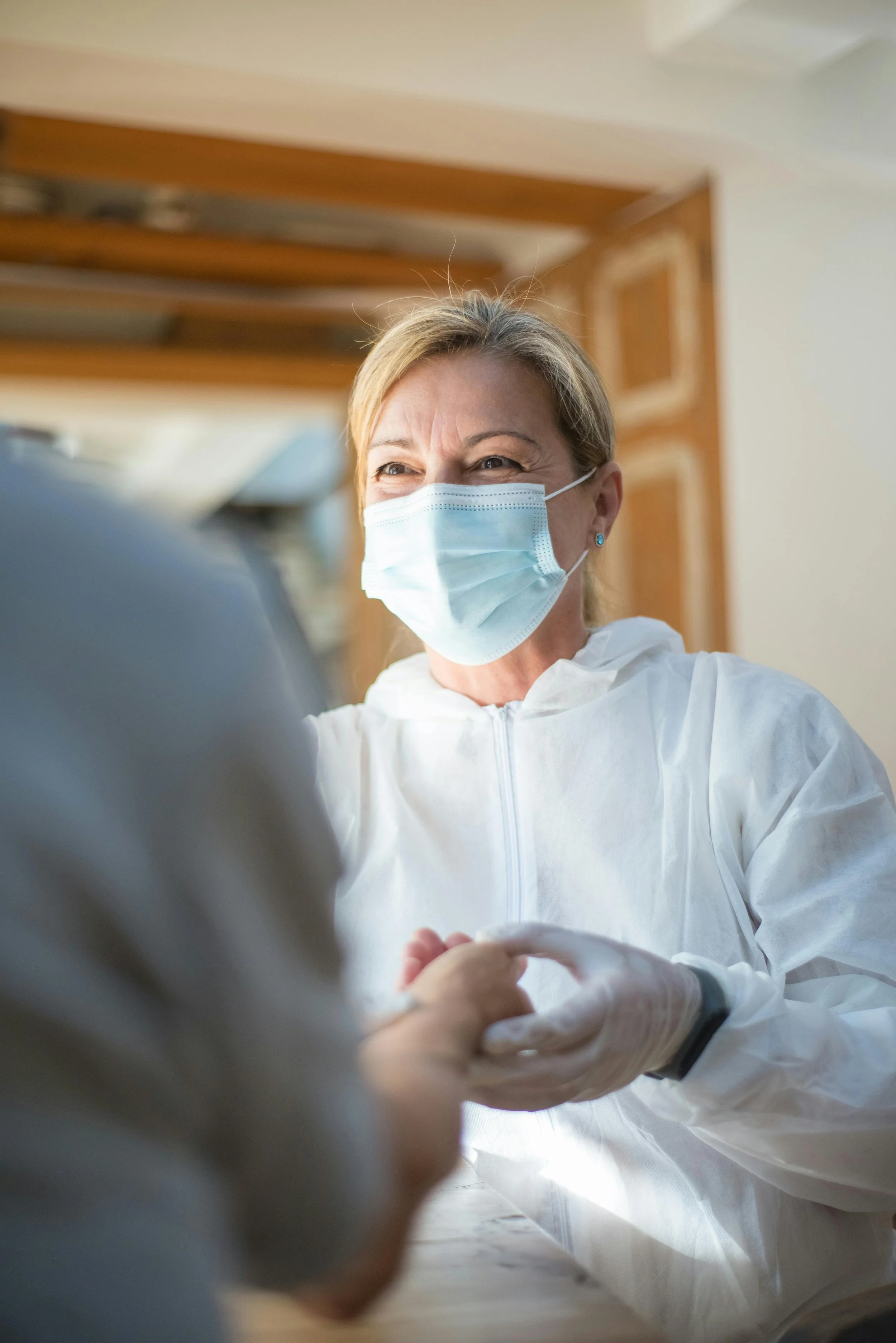 A woman wearing a protective face mask and gloves is holding hands with another person during a medical or healthcare setting, smiling with her eyes.
