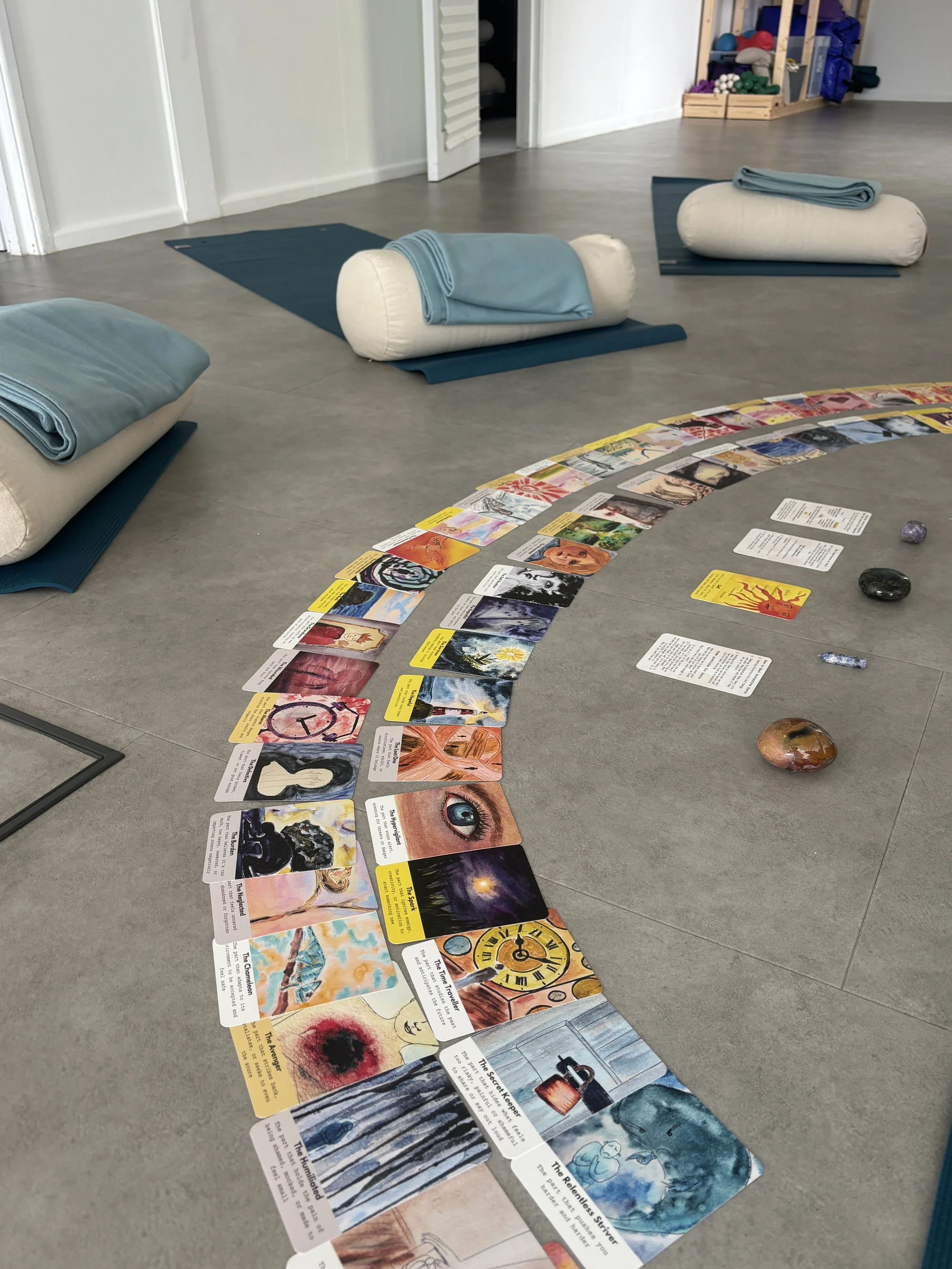 A room with three yoga mats and large rolled-up blankets, set up for an IFS breathwork session. In the foreground, there is a spiral-shaped arrangement of IFS cards with colorful illustrations and text, along with a few decorative crystals.