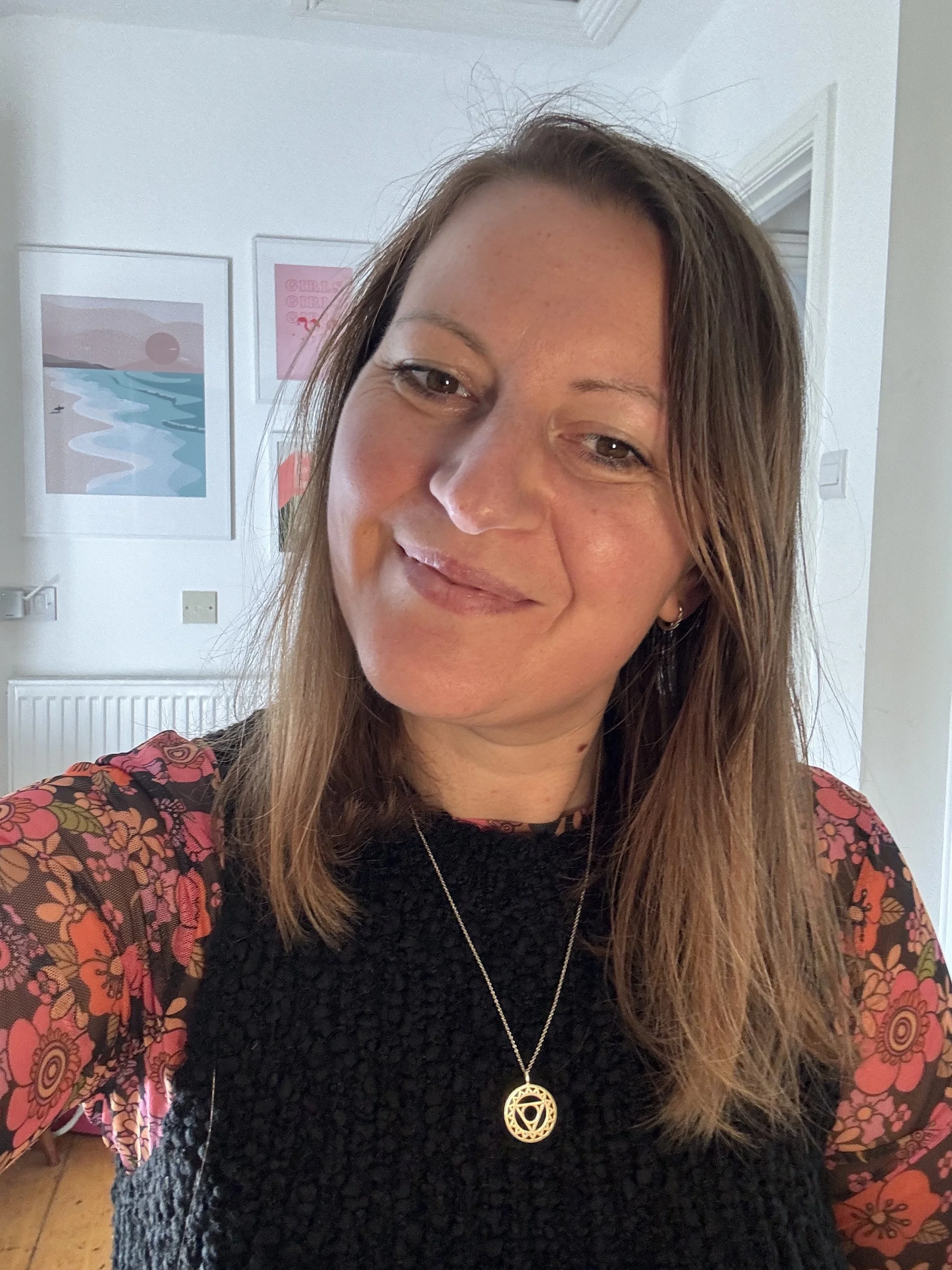 Hayley Hildred, Breathwork Practitioner. She has shoulder-length light brown hair smiling, wearing a black top with sheer floral sleeves and a gold necklace, in a room with pastel-colored artwork on the wall.
