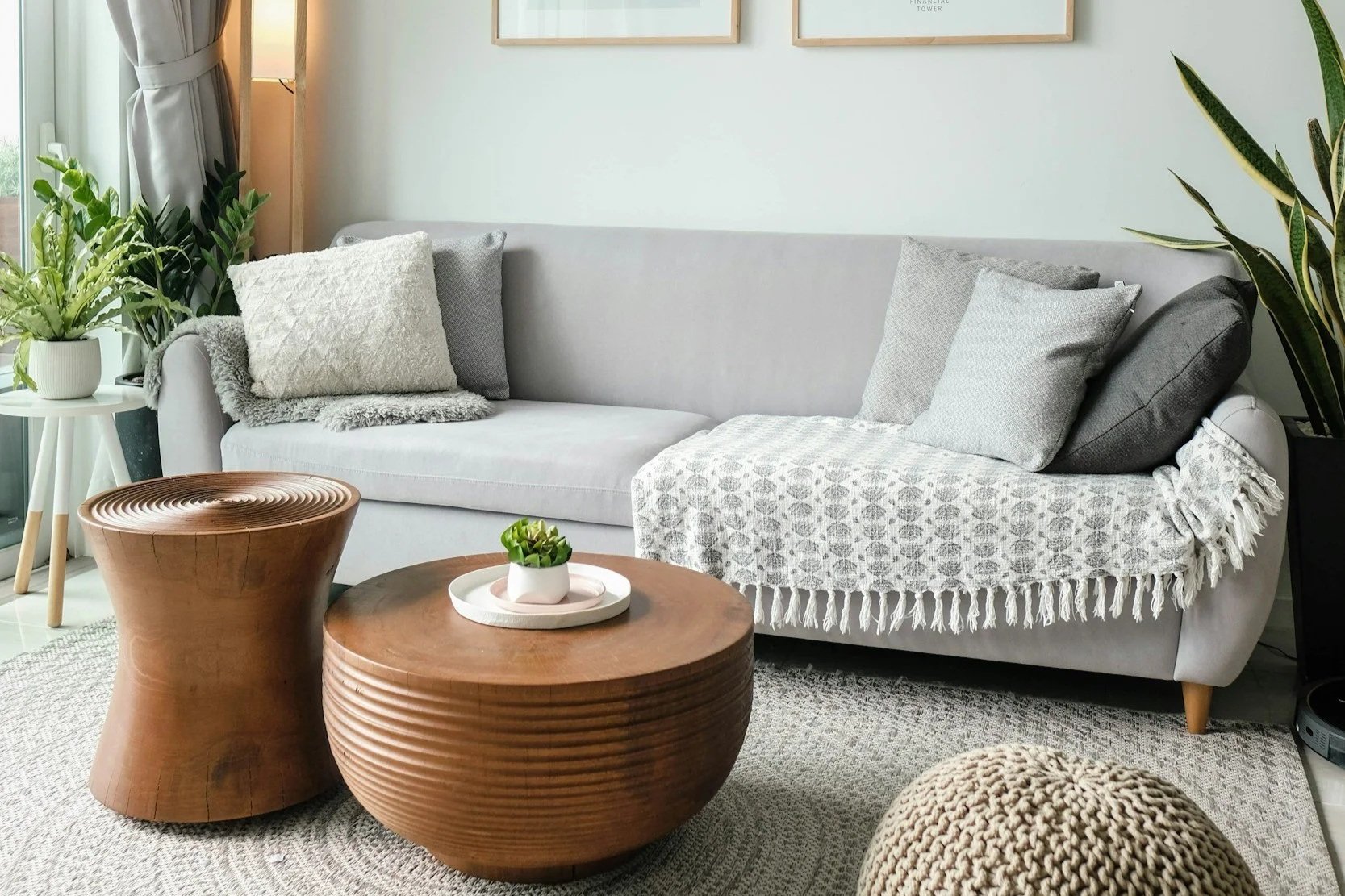 Cosy Therapy Space in Cornwall. Picture features a grey sofa, multiple throw pillows, a white blanket, a round wooden coffee table with a small plant, a textured pouf, and green indoor plants near a window with curtains.