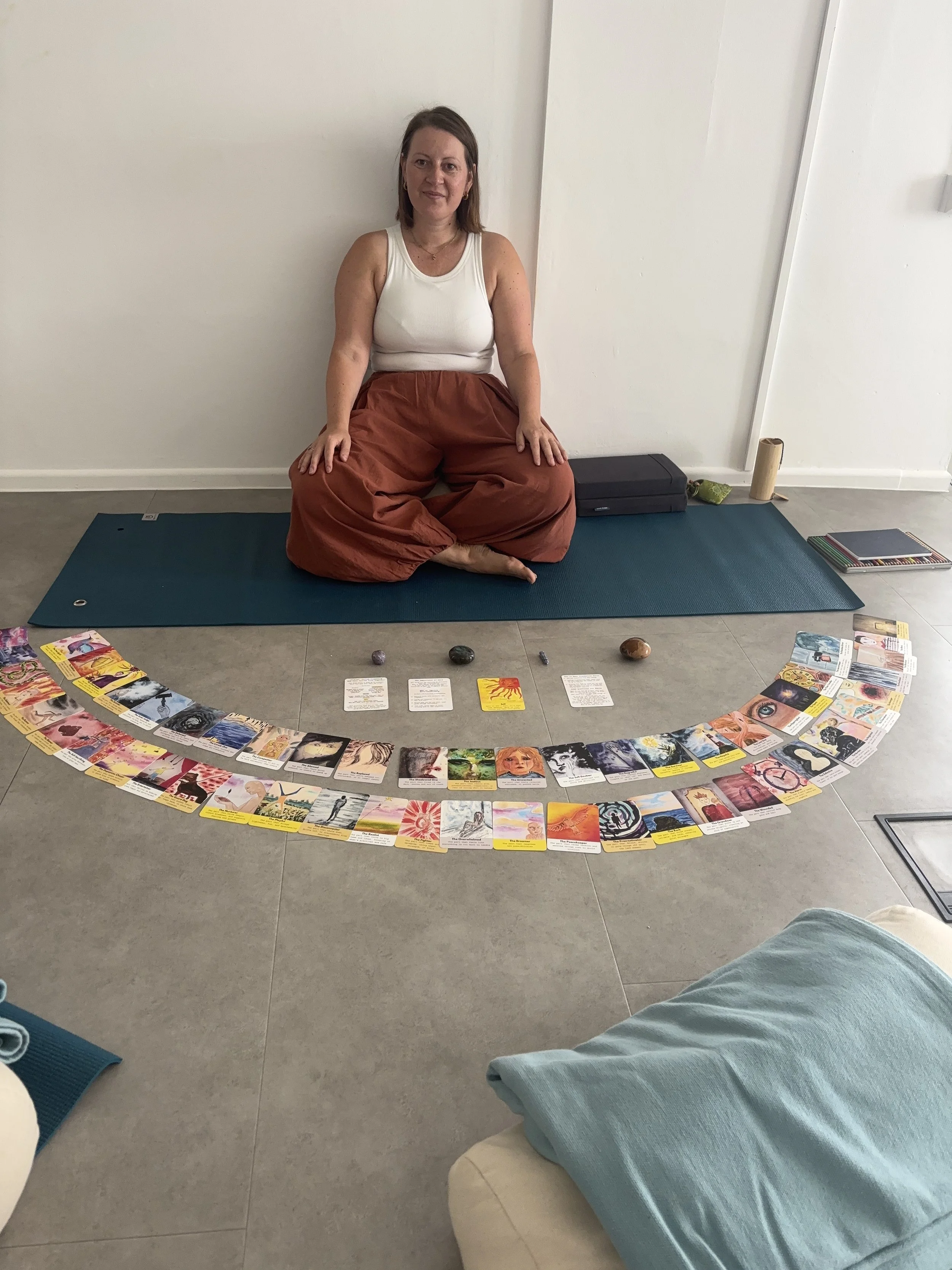 Person sitting cross-legged on a yoga mat with a circle of tarot or oracle cards laid out before them. The person is wearing a white tank top and rust-colored loose pants, with a black case and other items on the floor nearby.