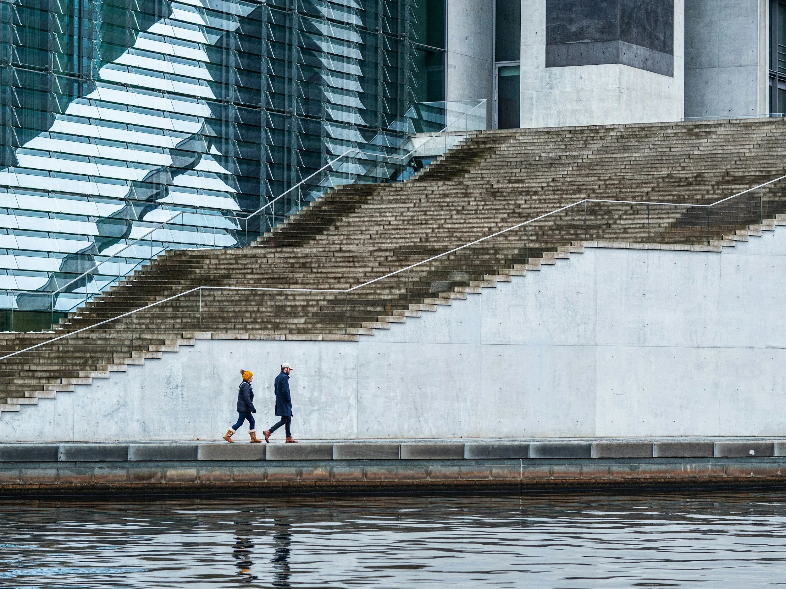Two people walking along a waterfront with stairs and a modern building in the background.