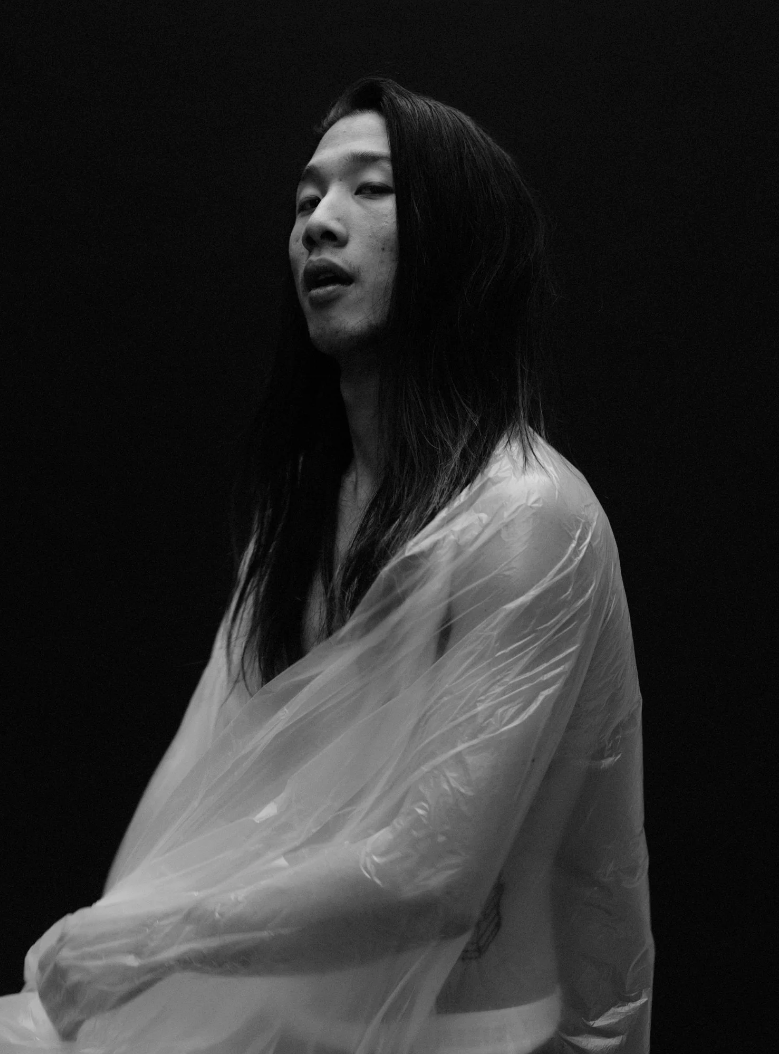 A black and white portrait of a person with long dark hair, slightly open mouth, wearing a translucent plastic jacket against a dark background.