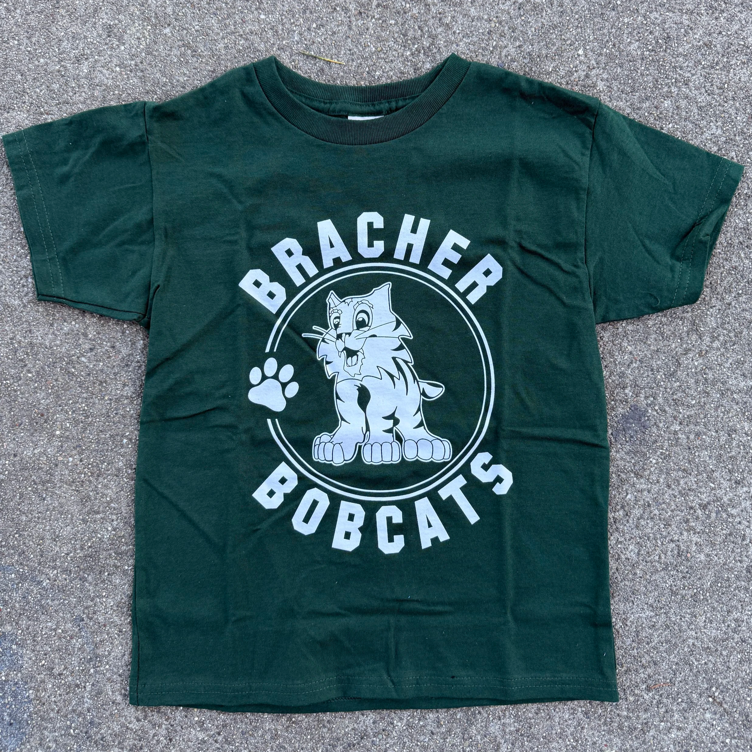 Green T-shirt with a cartoon bobcat and text 'Bracher Bobcats' and a paw print on a concrete surface.