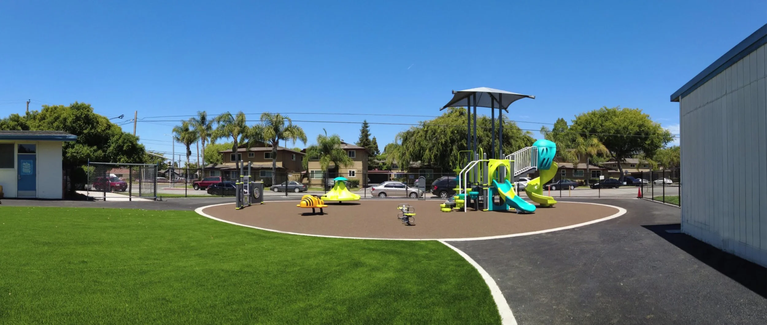 Bracher's playground area with slides and play structure surrounded by green grass, with trees and residential houses in the background under a clear blue sky.