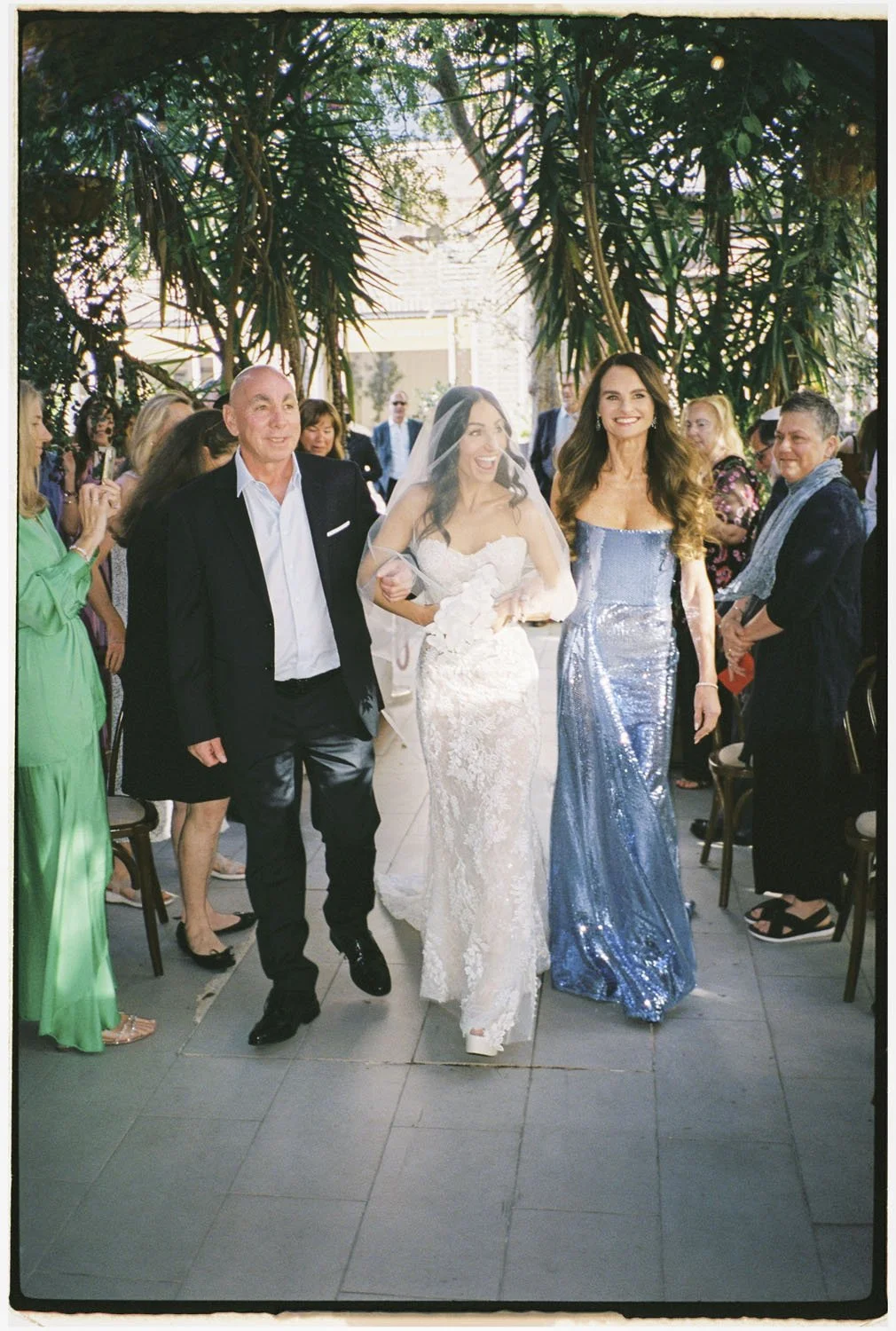 A bride in a white lace wedding dress walking down the aisle with two people, surrounded by guests in colorful attire, outside under a canopy of green trees, during a wedding celebration.