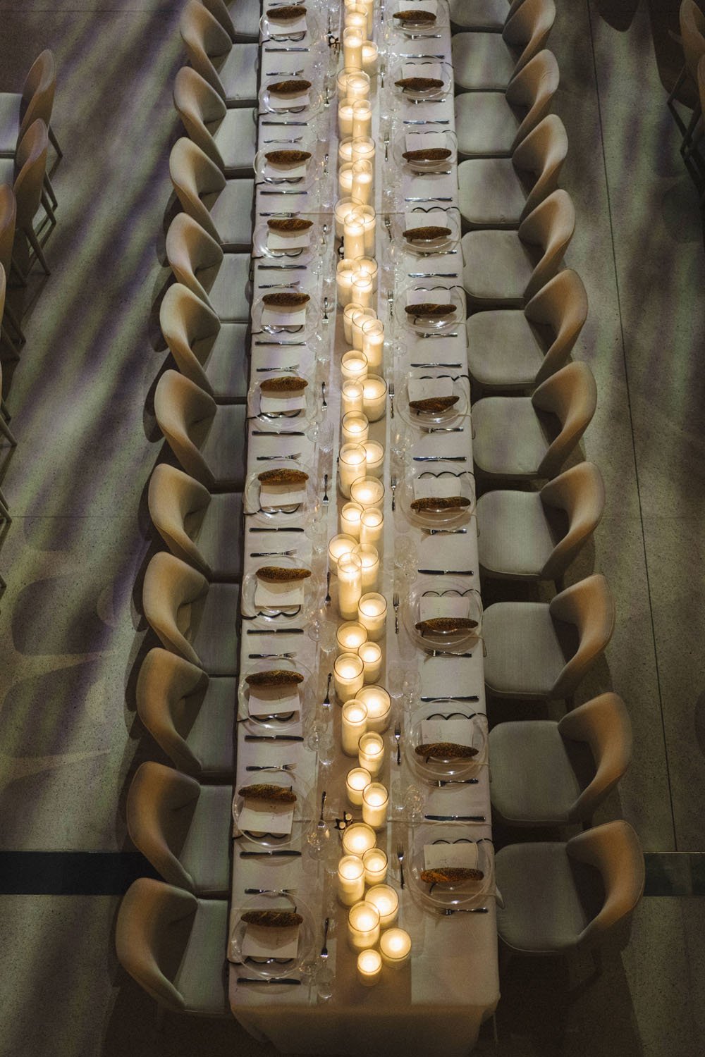 Long dining table set for a formal dinner with white tablecloths, candles, plates, silverware, and glasses, surrounded by beige chairs.