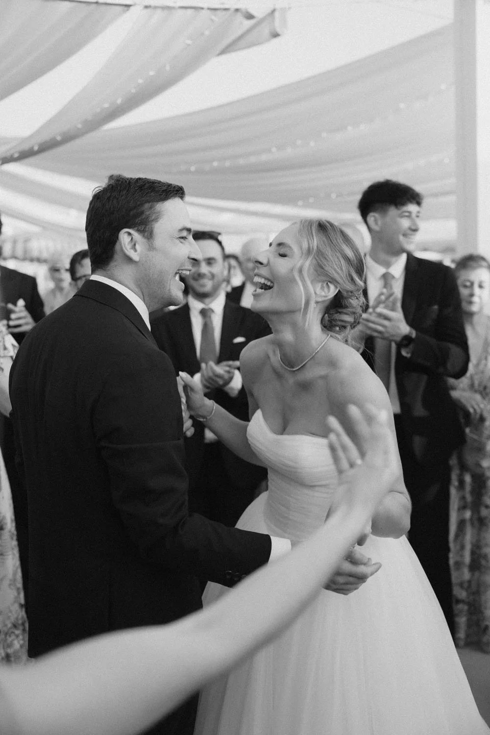 A bride and groom laughing and dancing at their wedding reception with friends around them.