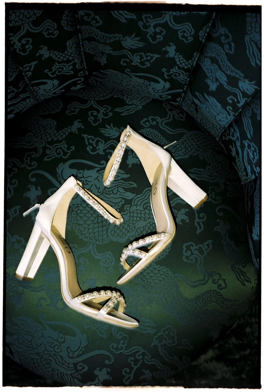 A pair of white high-heeled shoes with pearl decorations sit on a dark fabric surface with a dragon pattern.