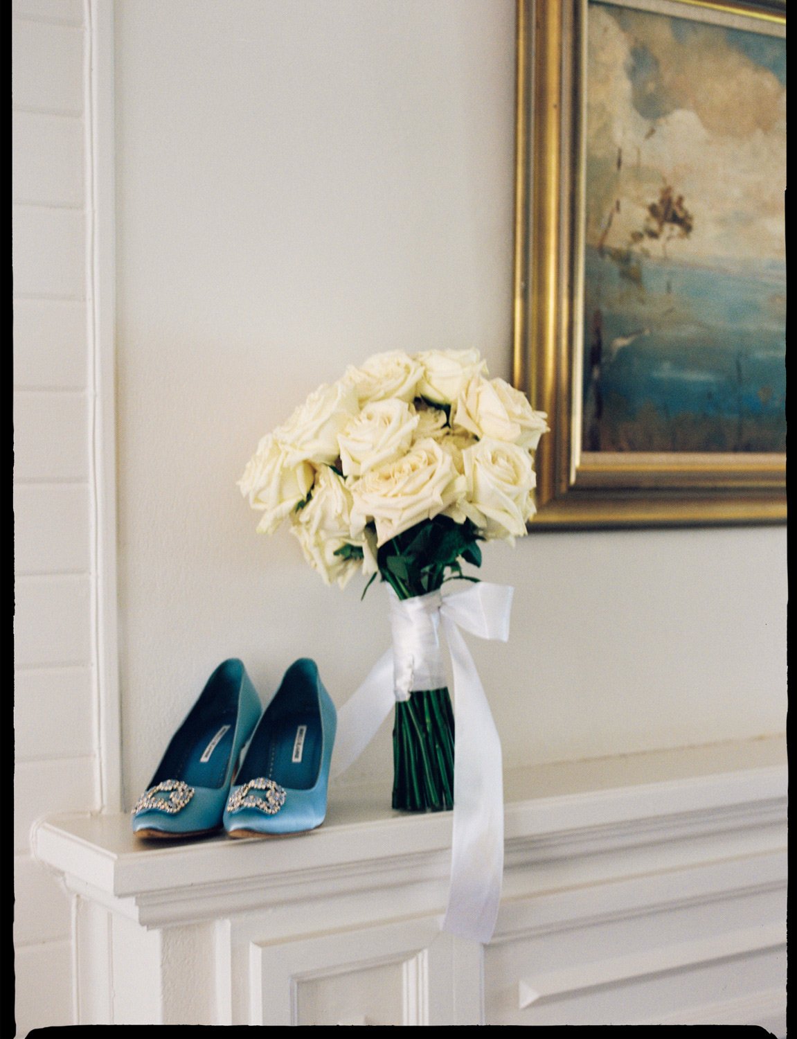 A pair of blue high-heeled shoes with decorative embellishments on the toes, a bouquet of white roses with green stems wrapped in white ribbon, and a framed painting hanging on a beige wall.