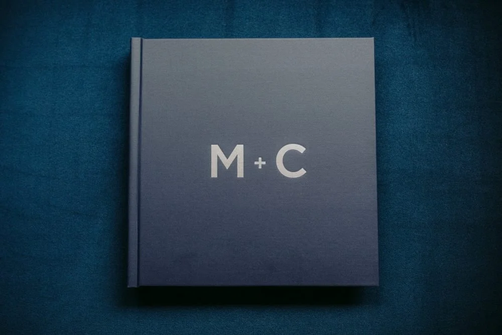 A hardcover book with a blue cover featuring the initials 'M + C' in white, placed on a blue fabric surface.