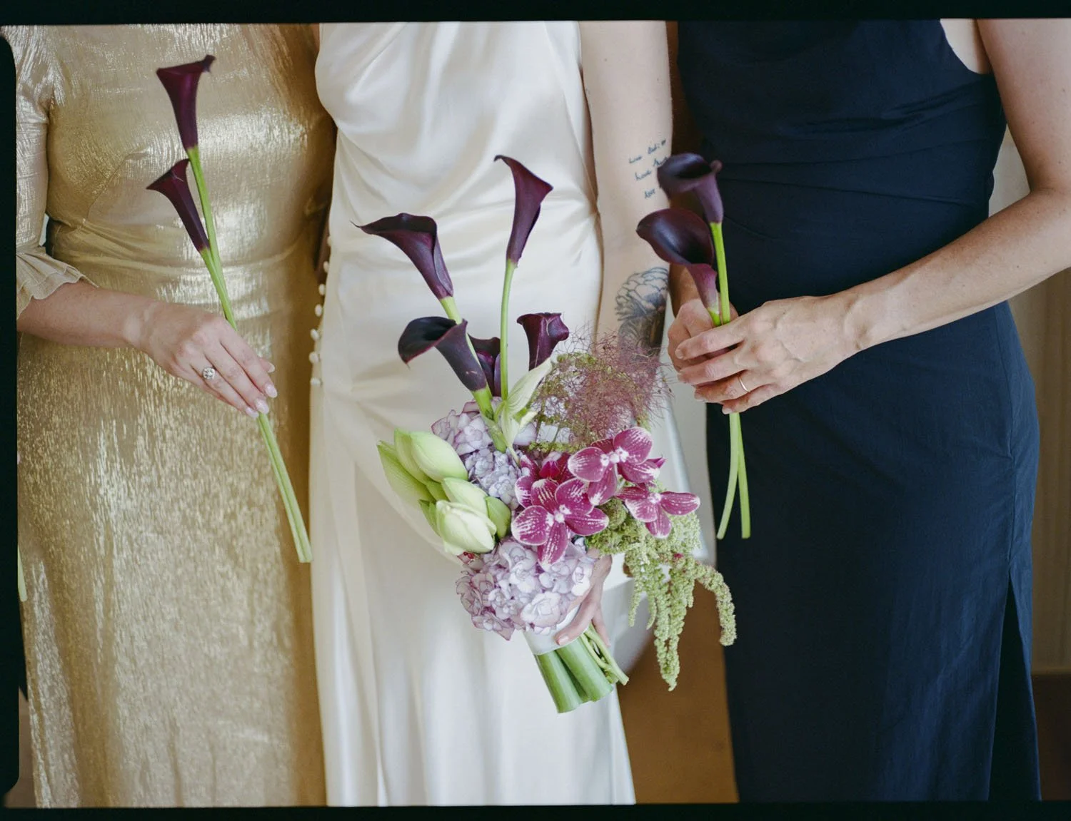 Three women standing together, holding a bouquet of flowers, including calla lilies, orchids, and greenery, dressed in formal attire.