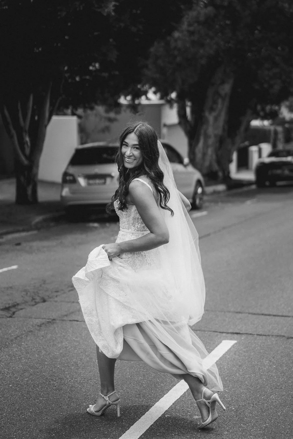 A woman in a wedding dress lifting her skirt and smiling while walking on a street lined with cars and trees.