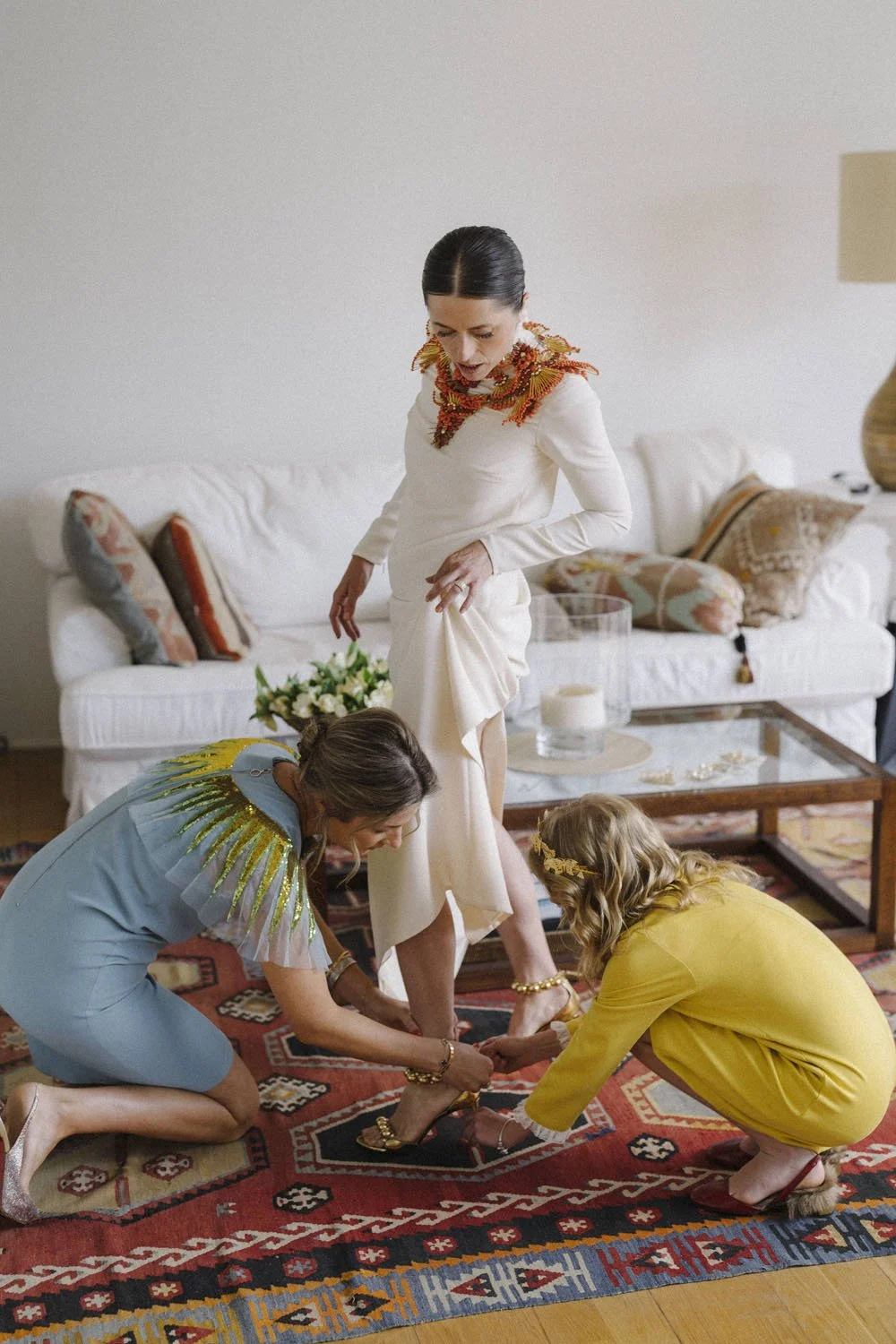 A woman dressed in white stands on a rug, while two women in colorful dresses assist her with her shoes in a living room.