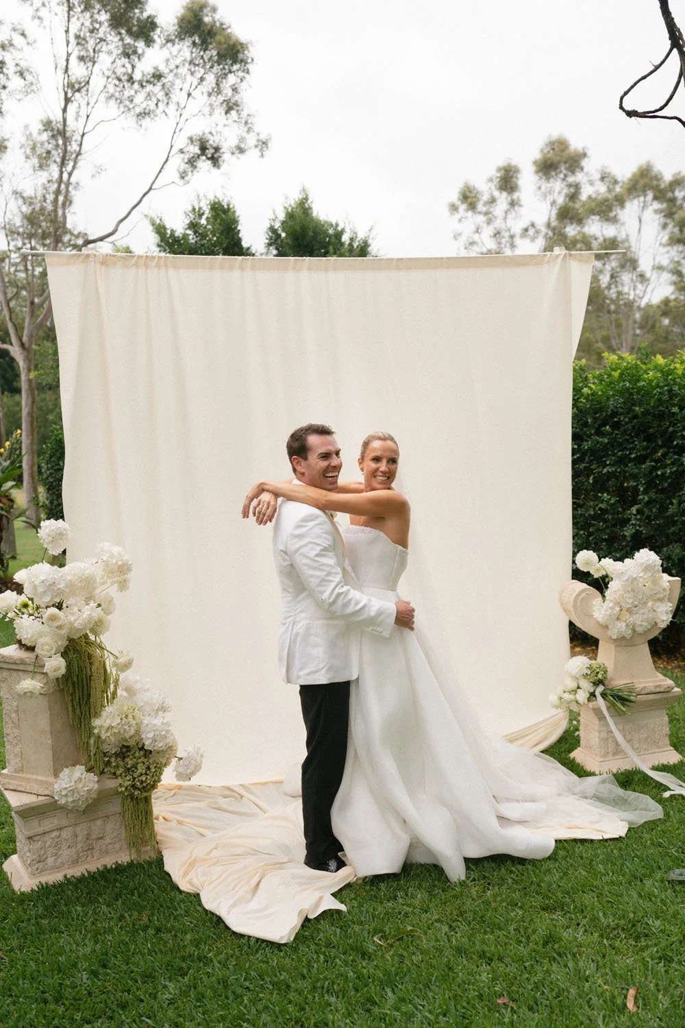 A bride and groom in wedding attire celebrating outdoors, standing in front of a cream-colored backdrop with flower arrangements on columns.