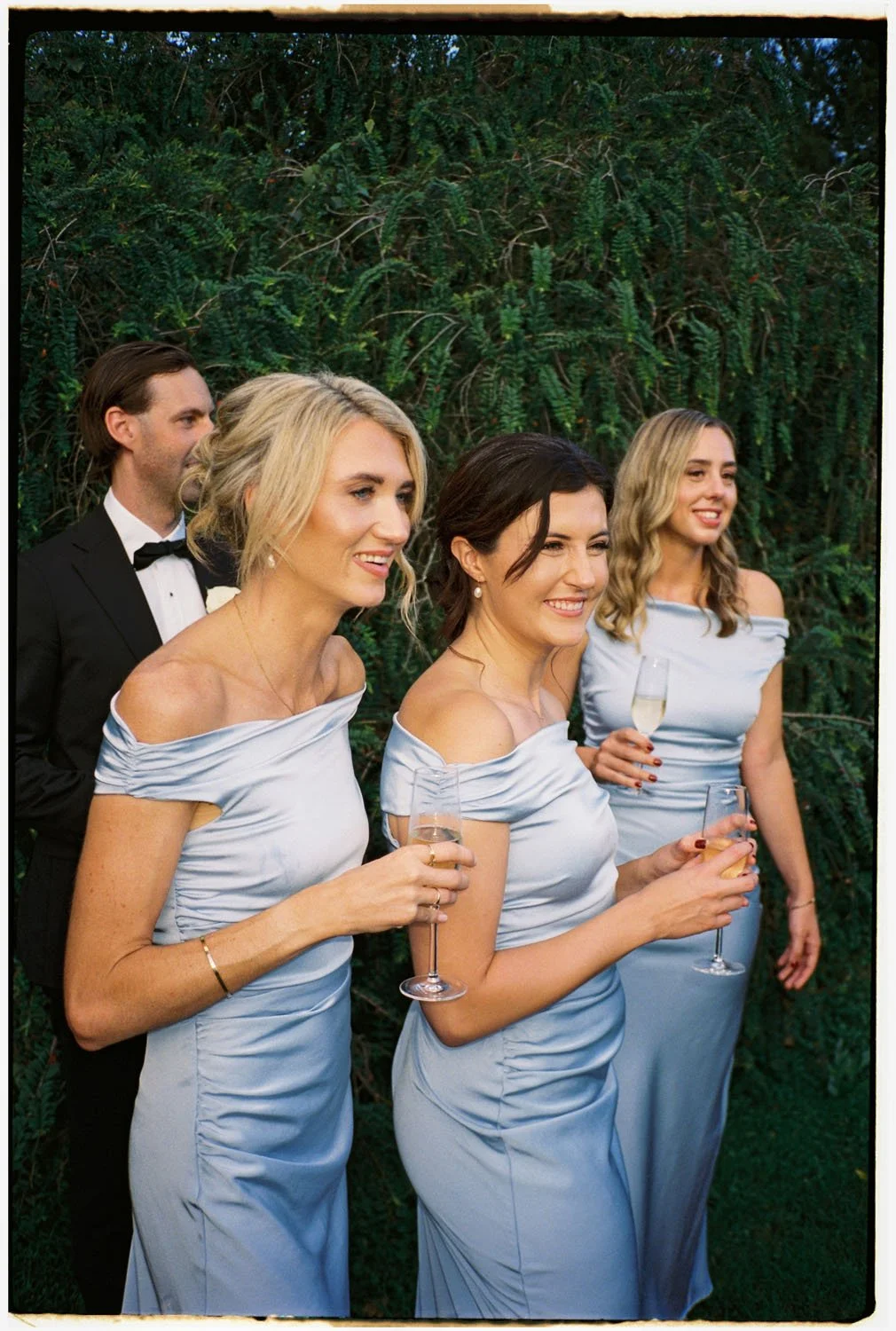 Group of four people, three women and a man, dressed in formal attire at an outdoor wedding or celebration, with greenery in the background. The women are wearing light blue dresses and holding champagne glasses, while the man is in a black tuxedo wi