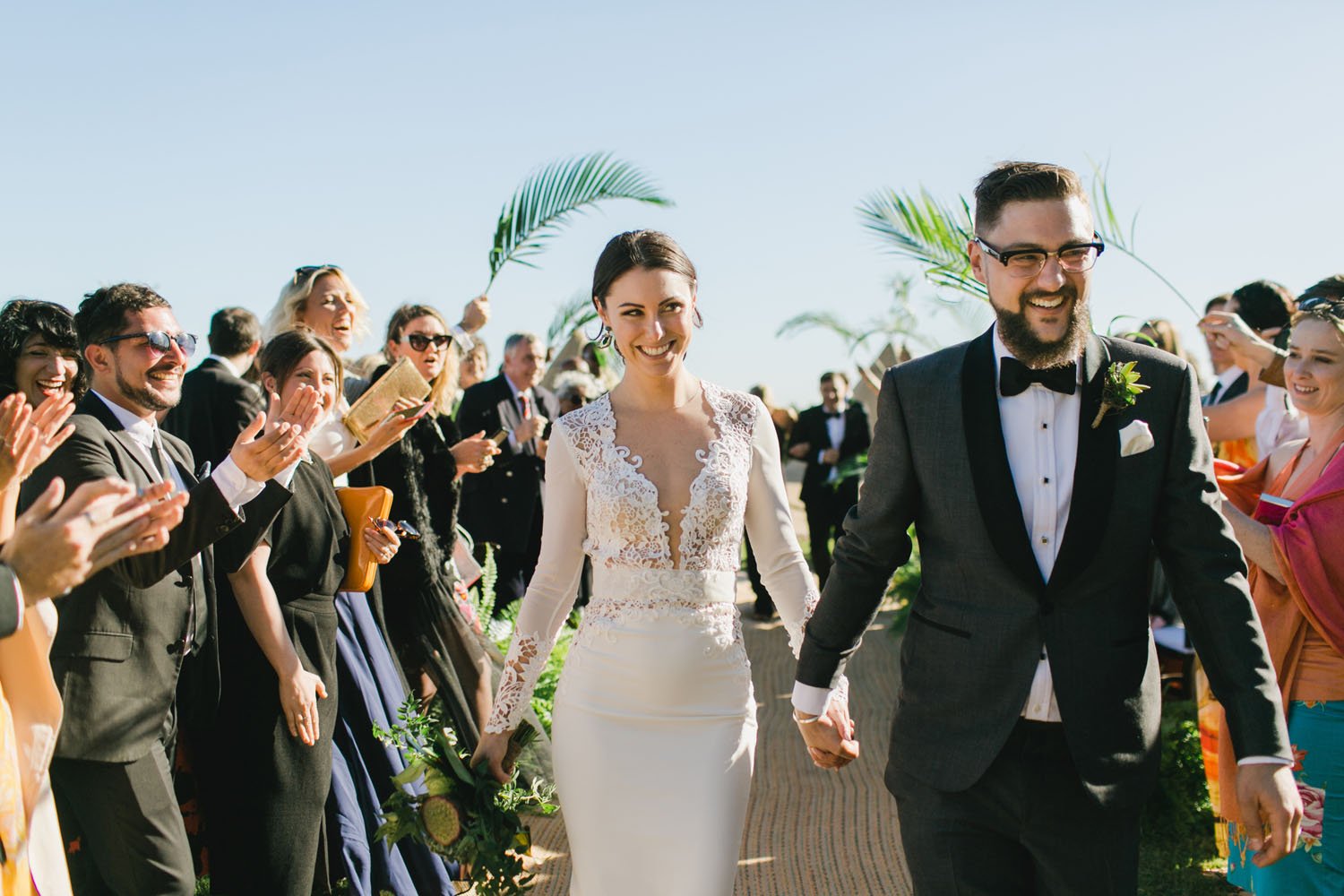 Bride and groom walking hand in hand on their wedding day, surrounded by cheering guests, outdoors on a sunny day with clear blue sky and palm leaves.