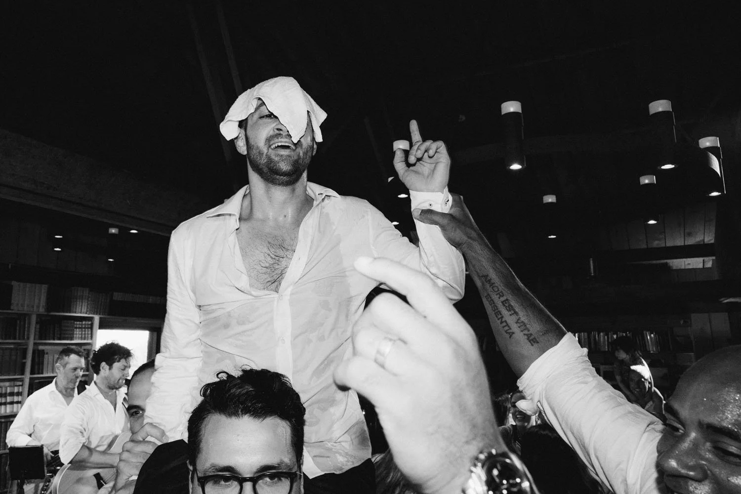 A man with a towel on his head, shirt open, and arm raised, is standing among a group of people in a room with bookshelves. Some people are reaching towards him, and he appears to be celebrating or jubilant.
