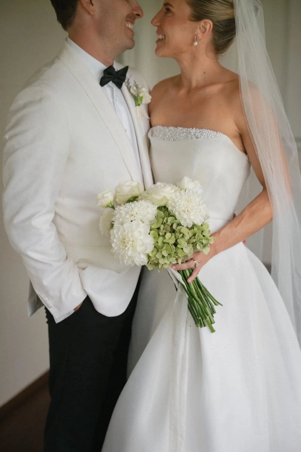 A bride and groom smile at each other, dressed in wedding attire, with the bride holding a bouquet of white flowers and greenery.