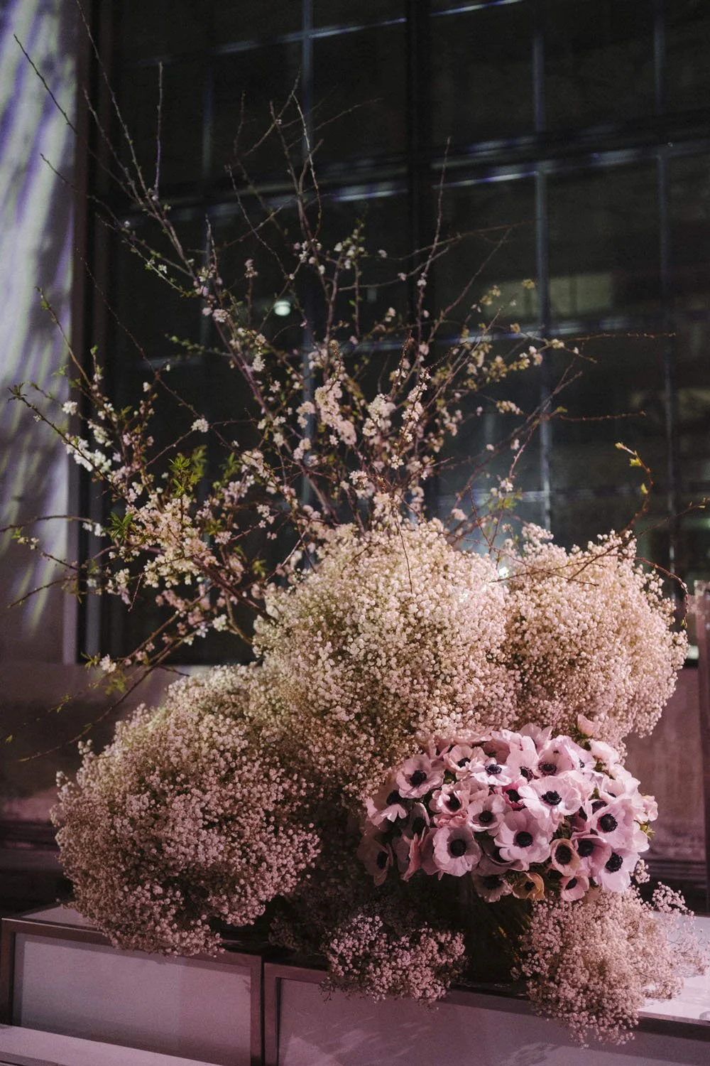 A large floral arrangement with pink cherry blossom branches, baby's breath, and pink anemone flowers on a white surface with a dark window in the background.