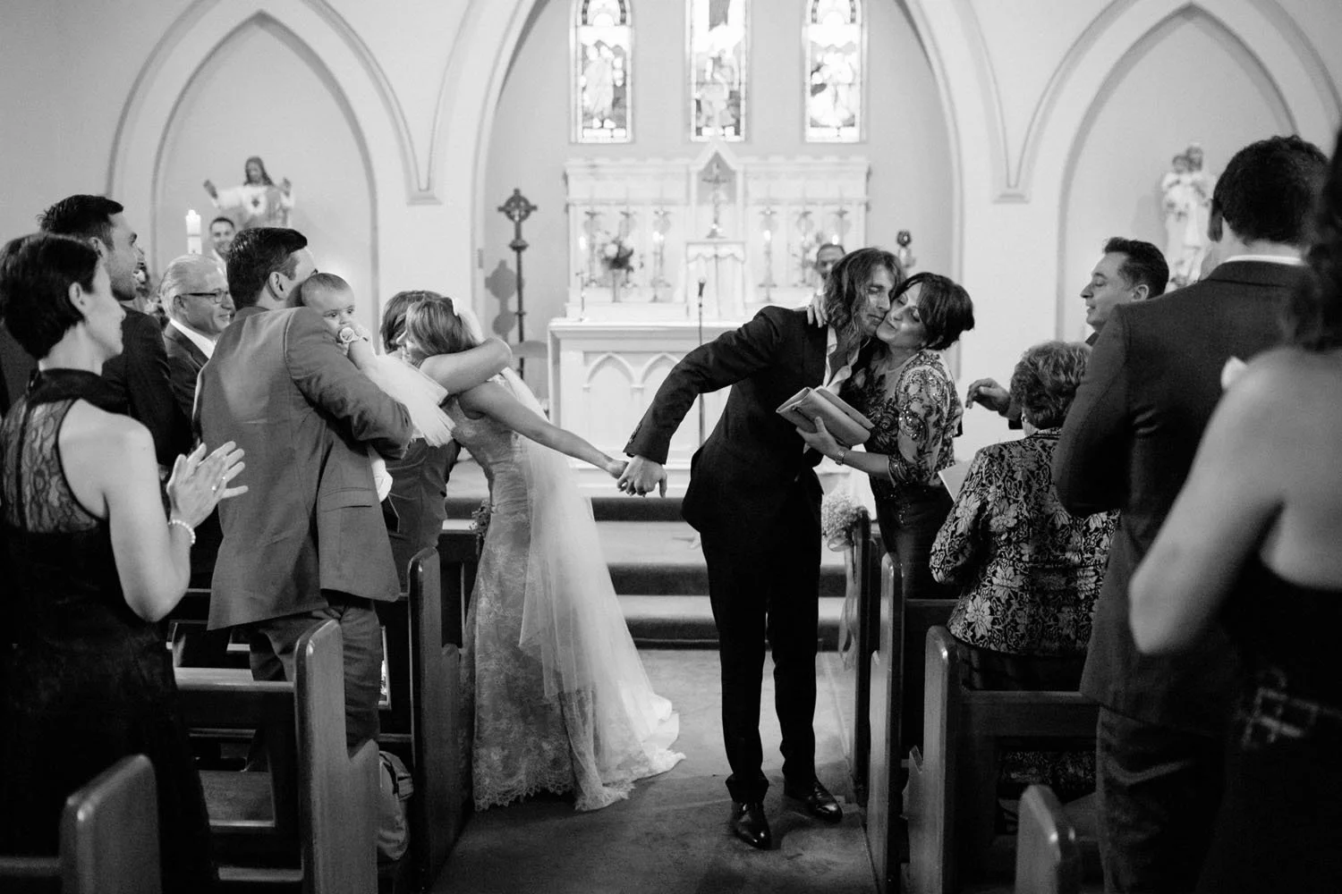 A black and white photo of a wedding ceremony inside a church, with a couple kissing at the altar and their wedding party pulling them apart, while guests watch and pray.